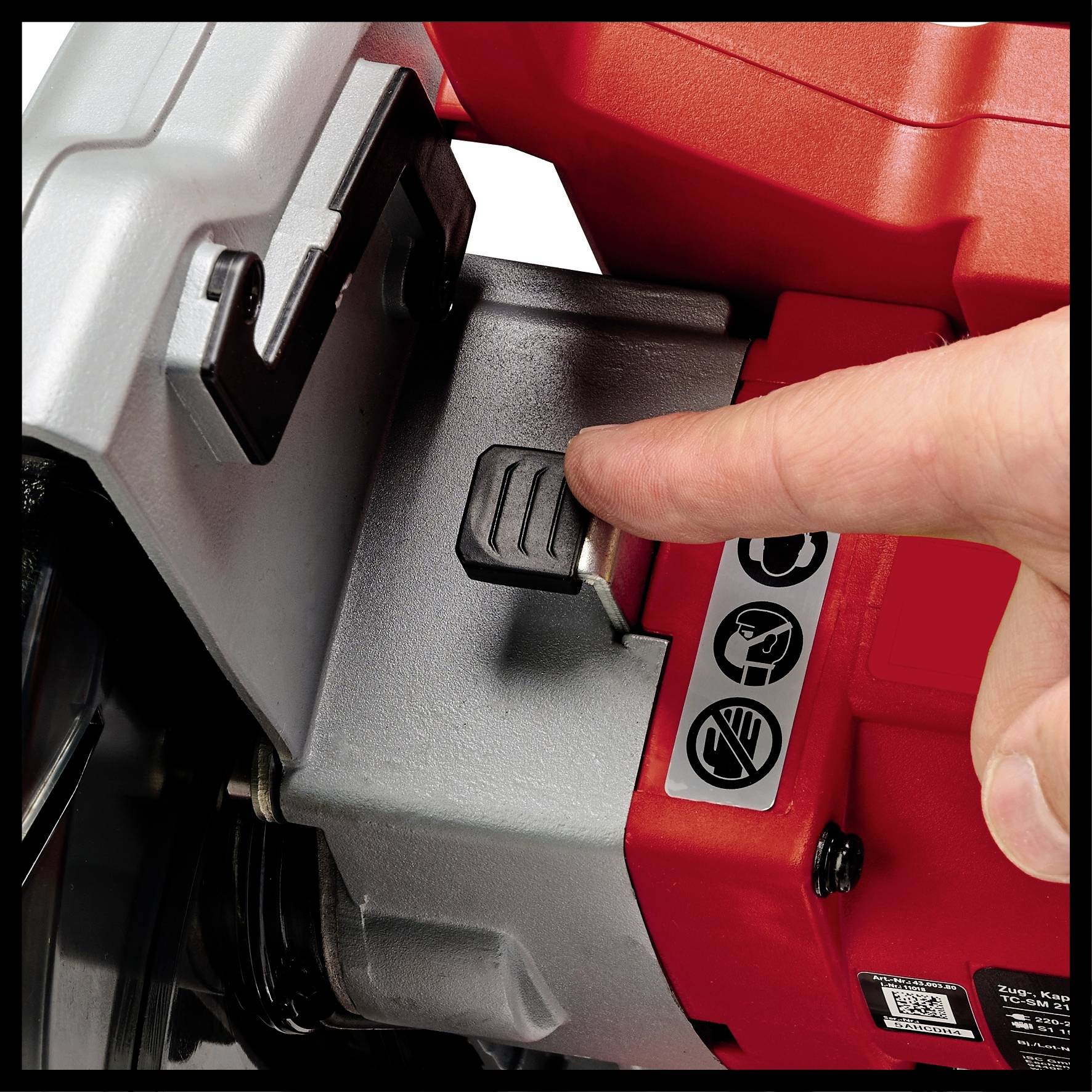 A hand is pressing a black lever on a red machine, with safety symbols nearby indicating protective measures.