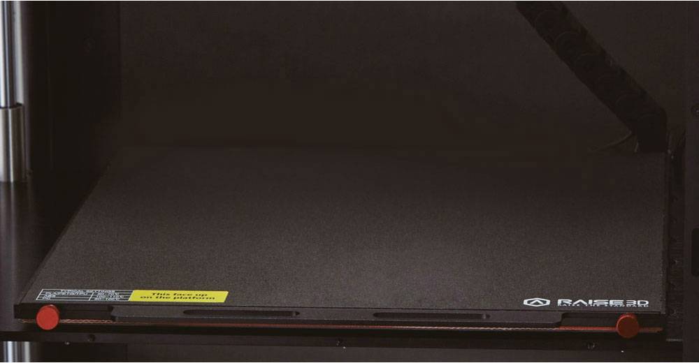 3D printer platform with a black surface. A yellow warning mark and two red fastenings are clearly visible.