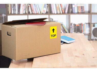 A brown cardboard box with a yellow 'TOP' sticker sits on a wooden table. Bookshelves are visible in the background.