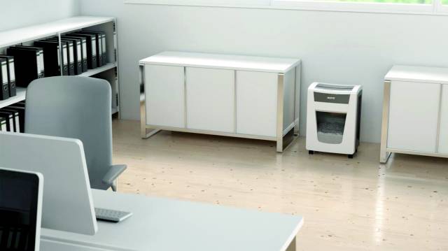 A bright office with wooden flooring, a desk, a bookshelf with files, and a shredder in the corner.