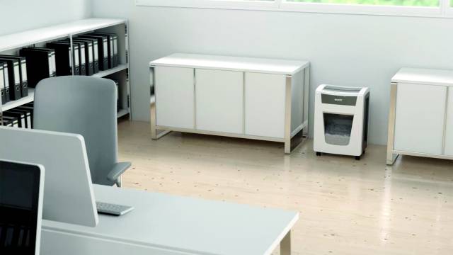 A modern office space with a document shredder on the floor. Desk with computer in the foreground, files on shelves.