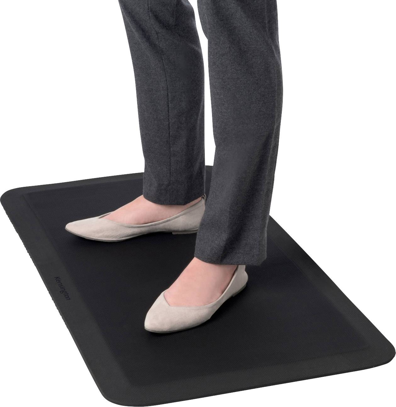 A person is standing on a black, rectangular mat, wearing grey trousers and beige shoes.