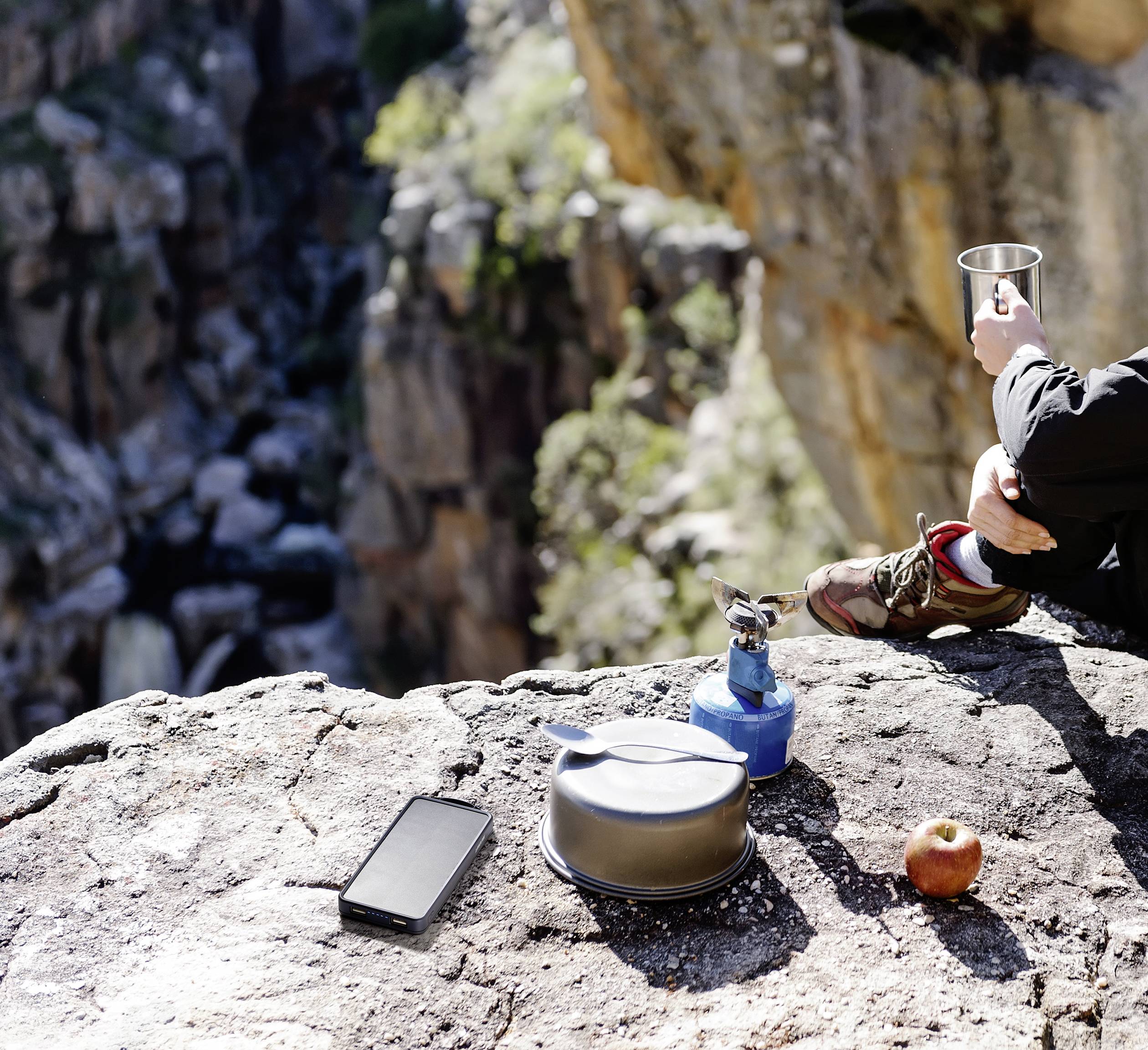 A person is sitting on a rock, holding a mug. On the rock, there is a camping stove with a pot, a smartphone, and an apple. In the background is a rocky gorge.