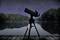 A telescope stands on a wooden deck, aligned towards a star-filled night sky over a tranquil lake with a wooded shoreline.
