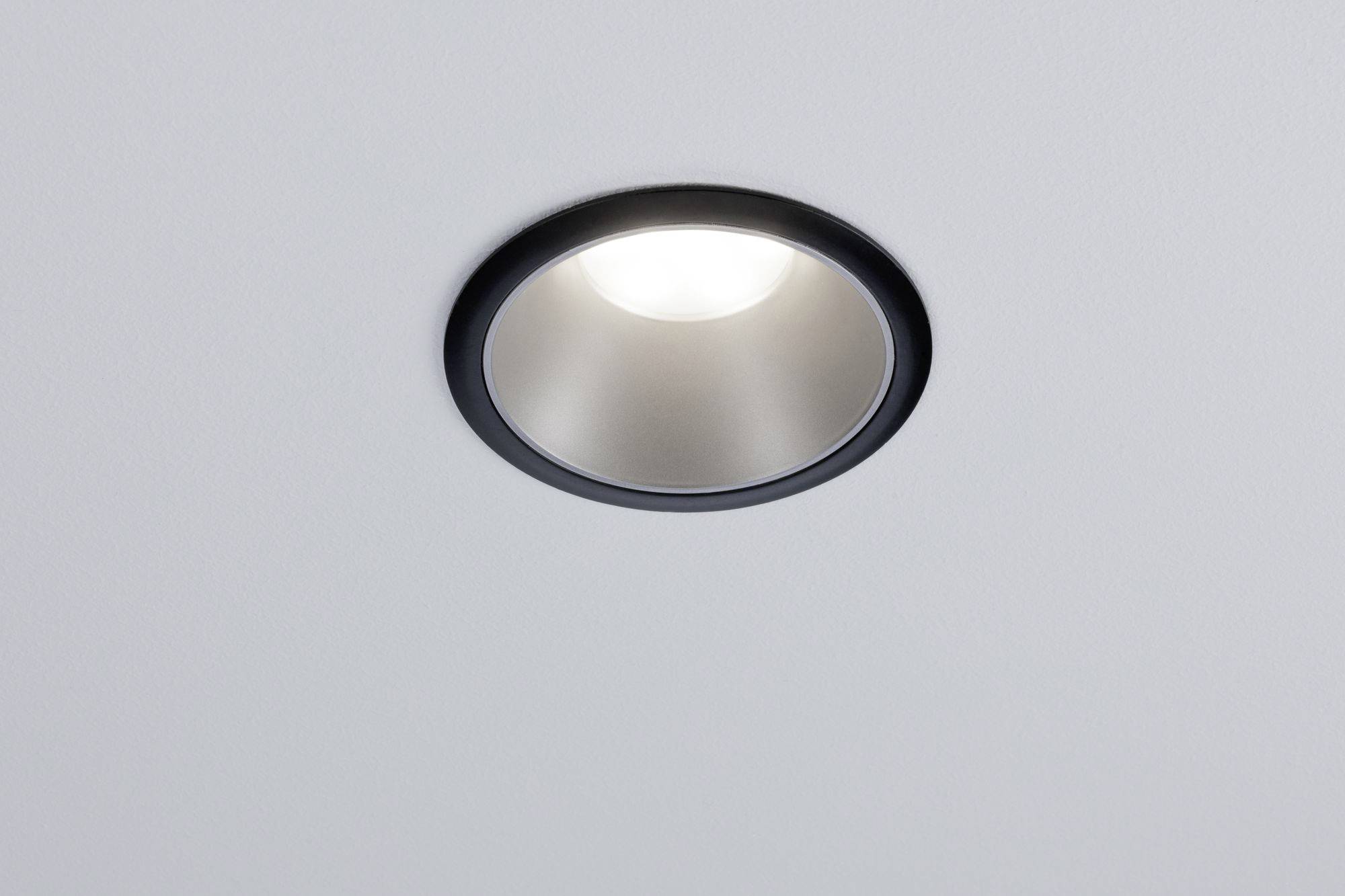 Ceiling light, round and silver with a black rim, brightly illuminating a white ceiling.