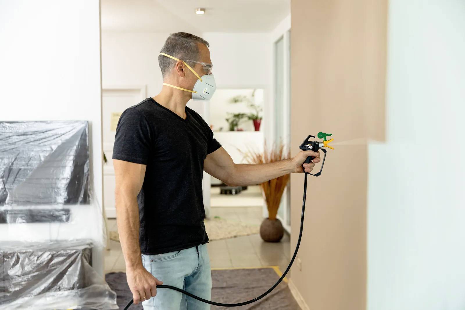 Man wearing a protective mask sprays paint on a wall, prepared with protective sheeting.