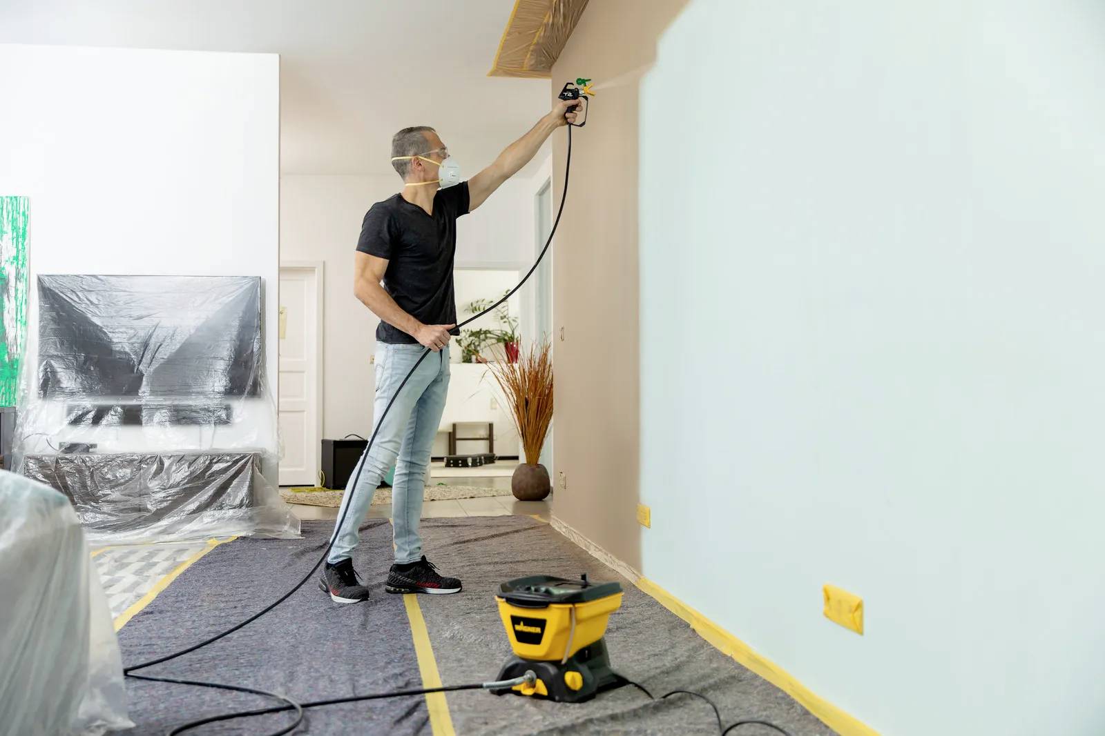 A man is spraying beige paint onto a living room wall using a paint spray gun. Furniture is covered with plastic sheeting, and the floor is protected.