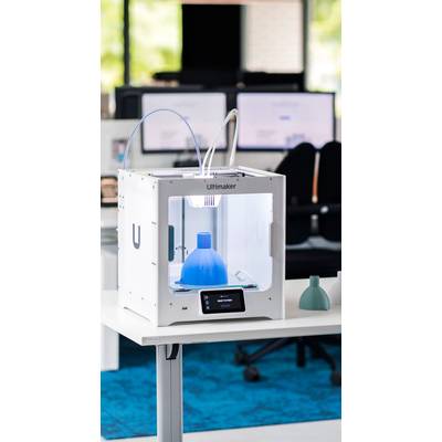 A 3D printer on a table is printing a blue object. Computer screens and office chairs are visible in the background.