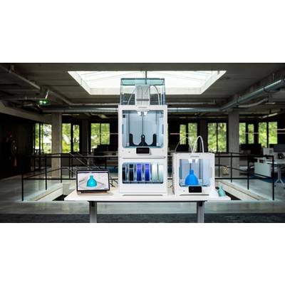 A modern office space with a desk featuring a laptop and several 3D printers. The laptop displays a blue 3D model.