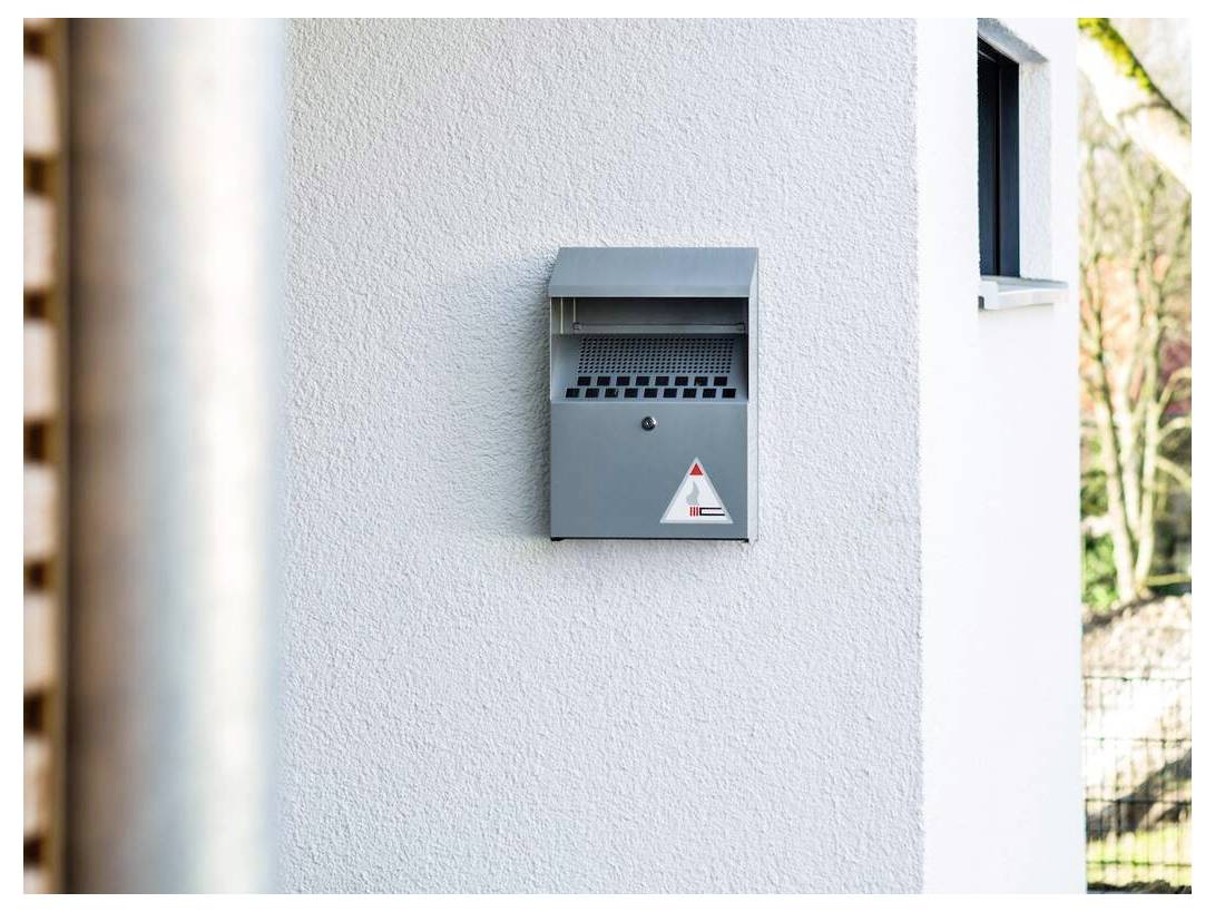 A gray mailbox is mounted on a white exterior wall of a building, partially shaded by sunlight.