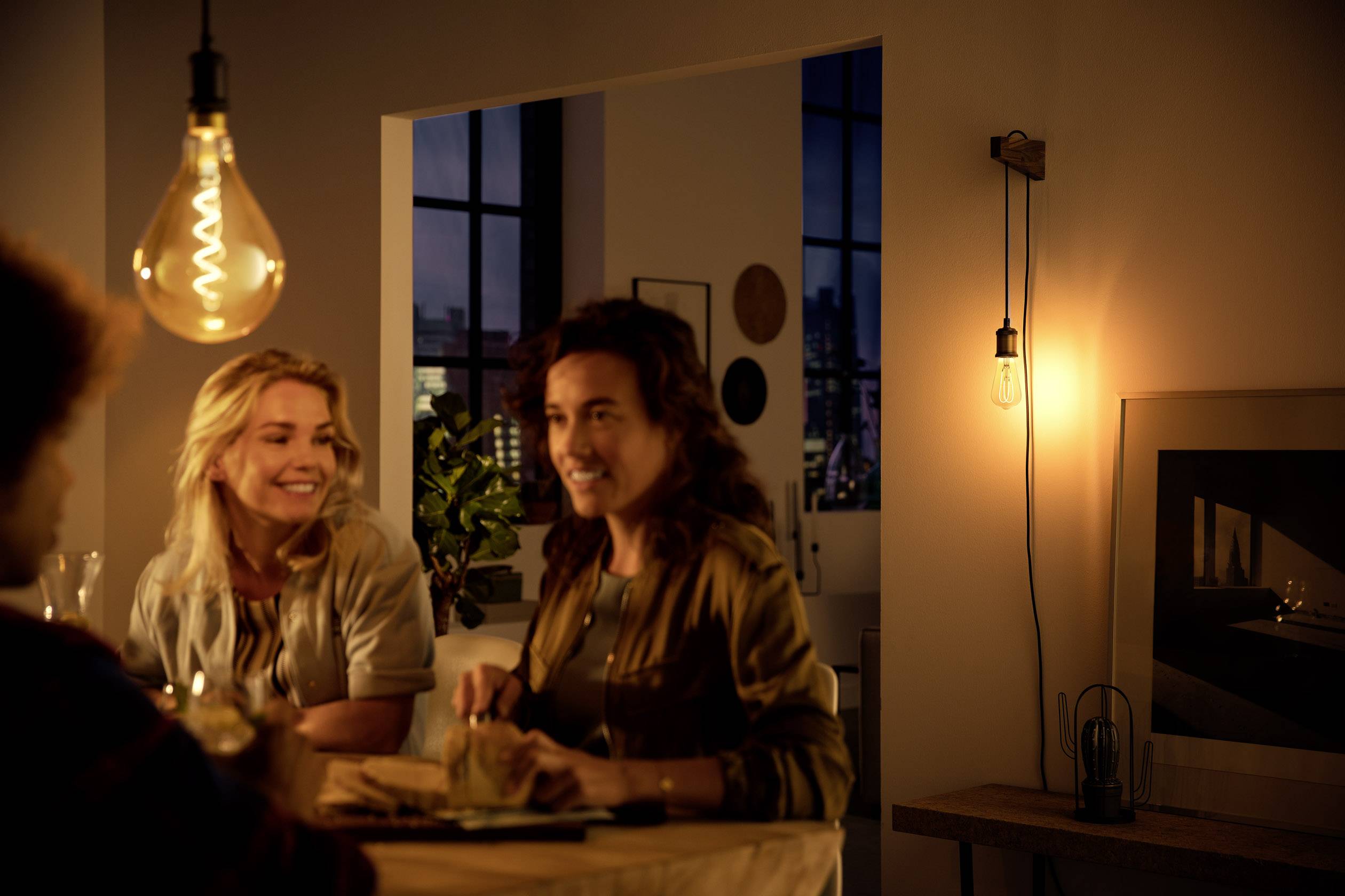 Three people are sitting at a table in an evening-lit flat with a cosy ambience. Large windows can be seen in the background.