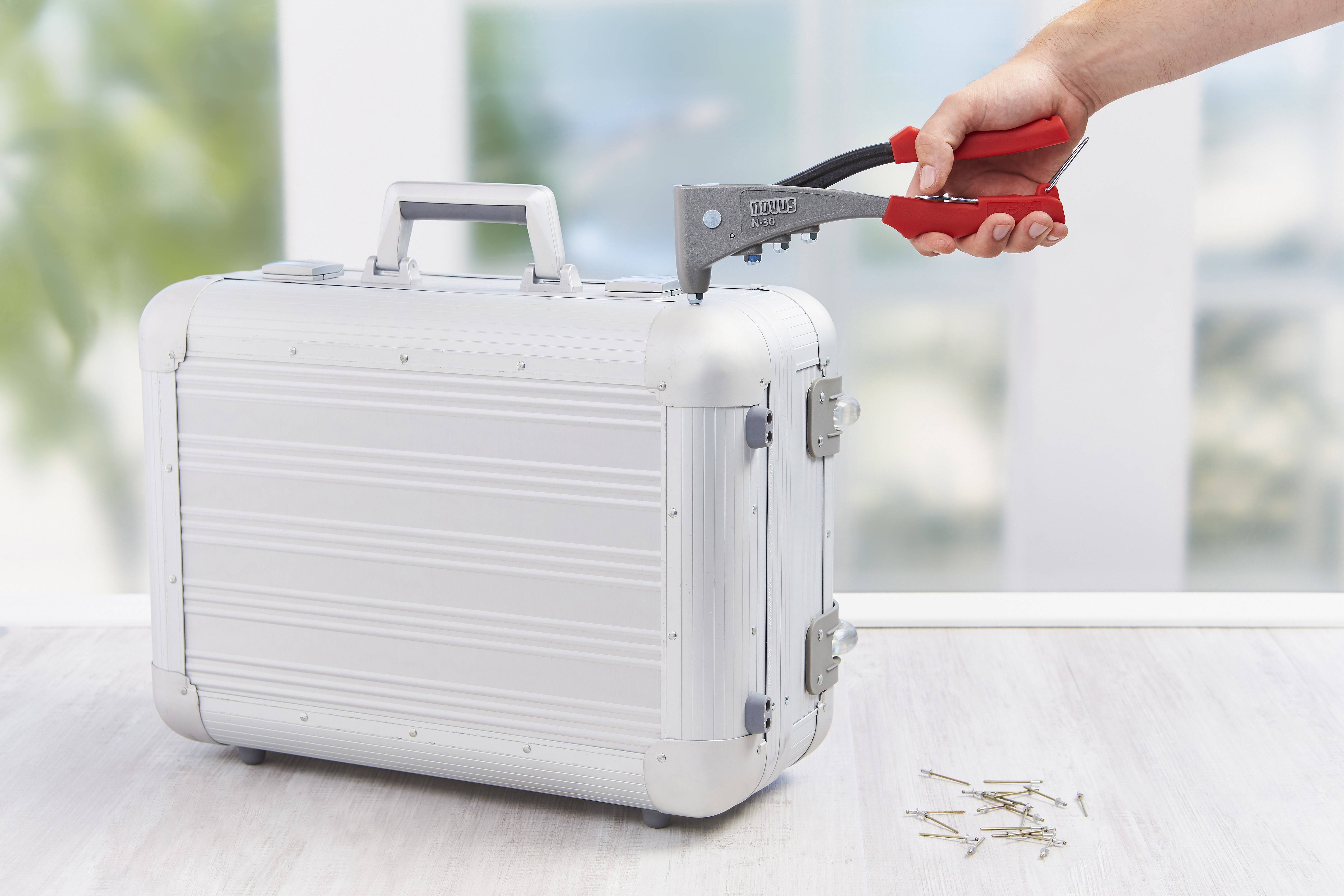 A hand holds a red riveting pliers over a silver metal case; several rivets are lying beside it on a table.