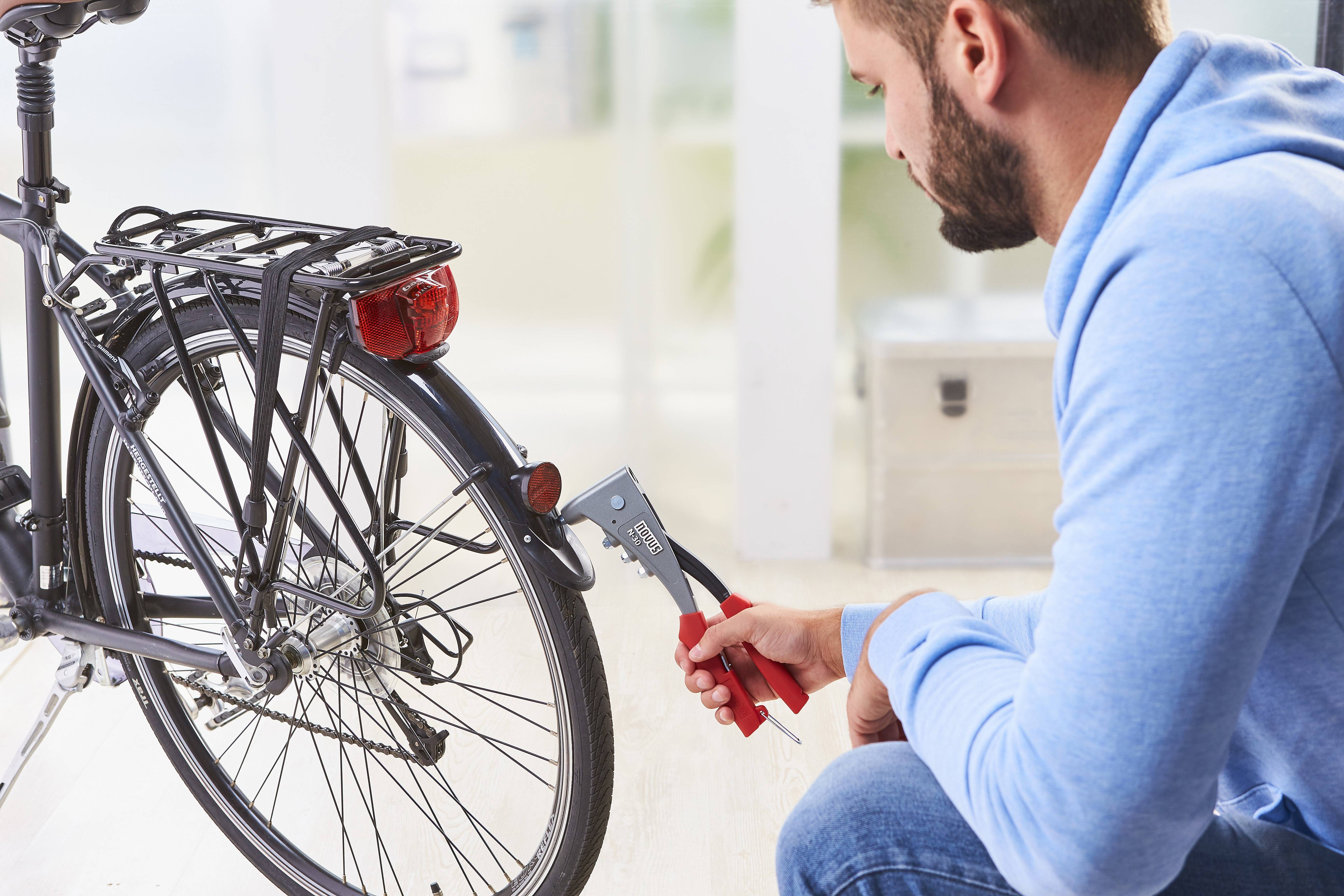 A man in a blue hoodie is repairing the rear wheel of a bicycle with a pair of pliers.