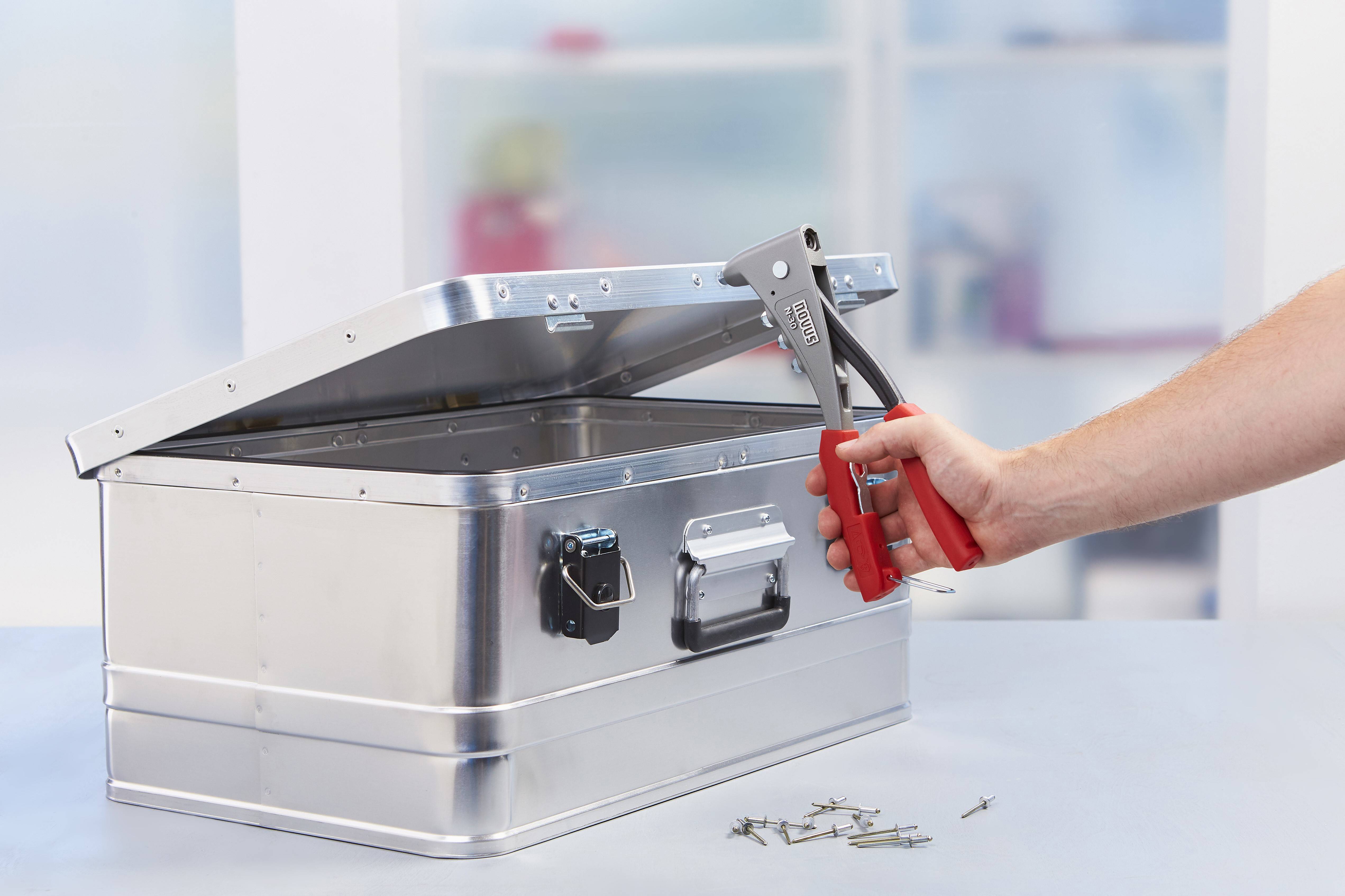 A hand holds a red rivet pliers over an open silver metal box, with loose rivets lying beside it on the table.