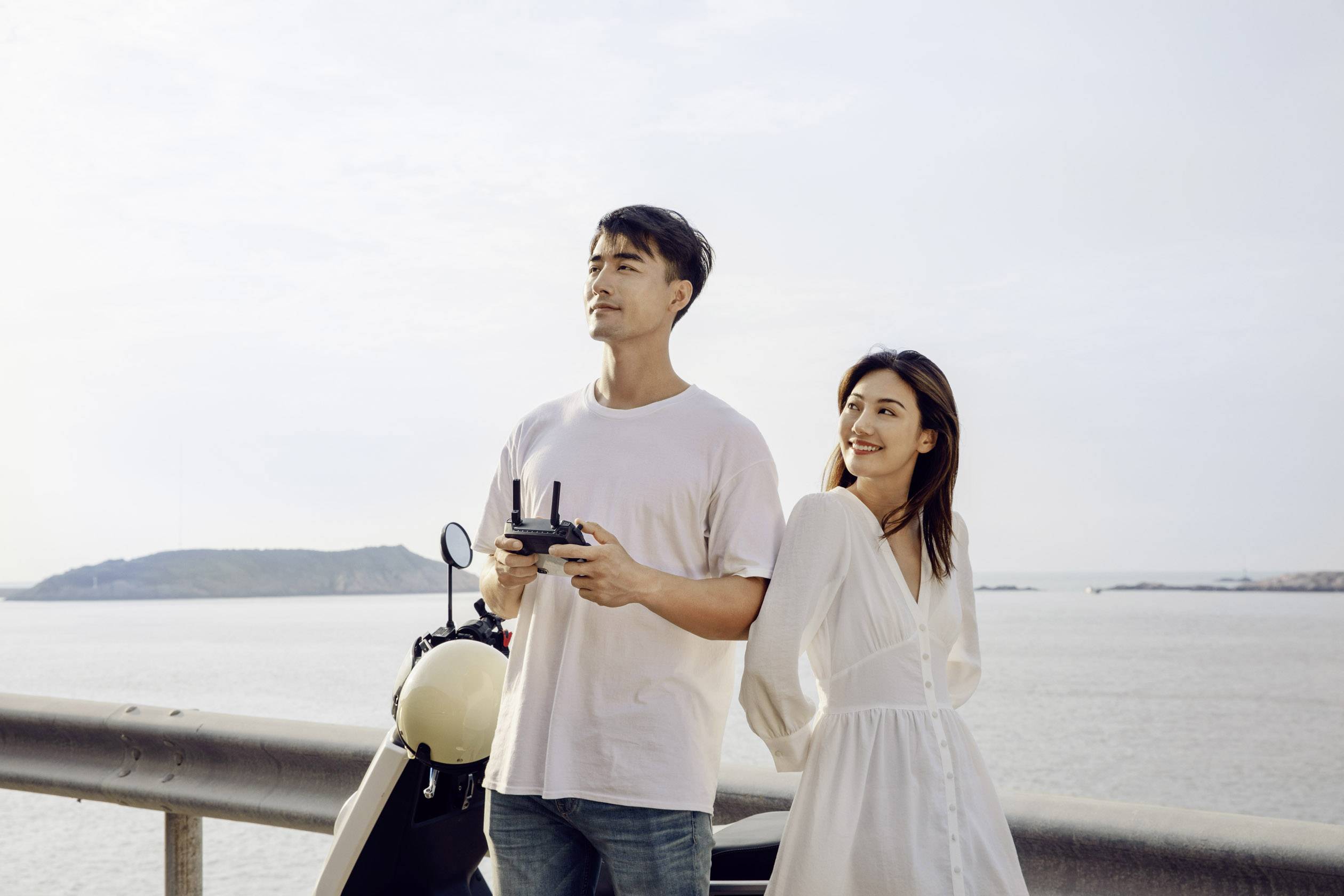 A man and a woman are standing by the sea, the man holding a remote control, while the woman smiles. A motorbike is parked beside them.