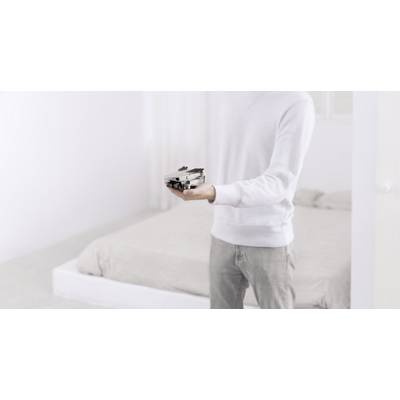 A person wearing a white jumper is holding a grey drone in their hand, standing in a bright bedroom in front of a bed.