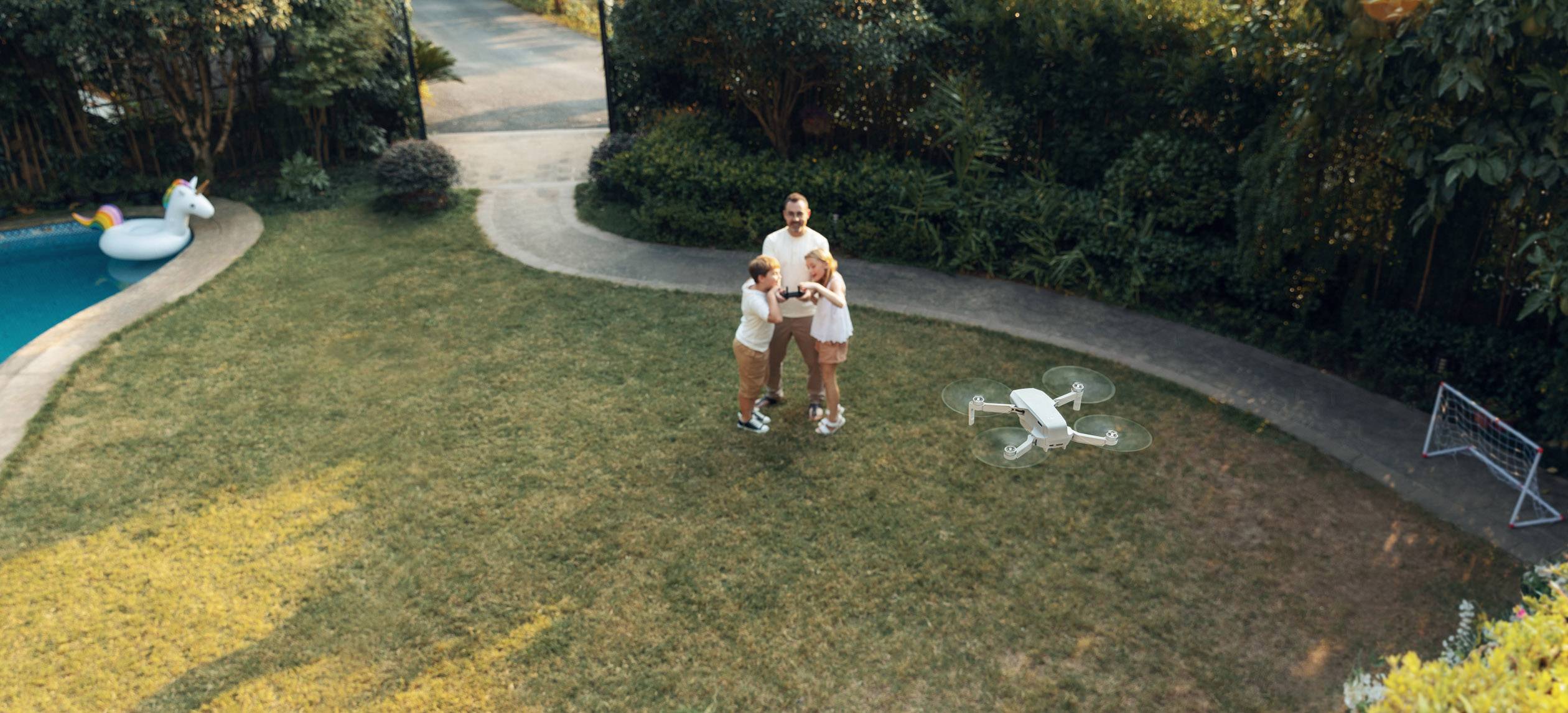 A group of three people are standing on a lawn, controlling a drone. Trees and a pool with an inflatable swimming animal can be seen in the background.
