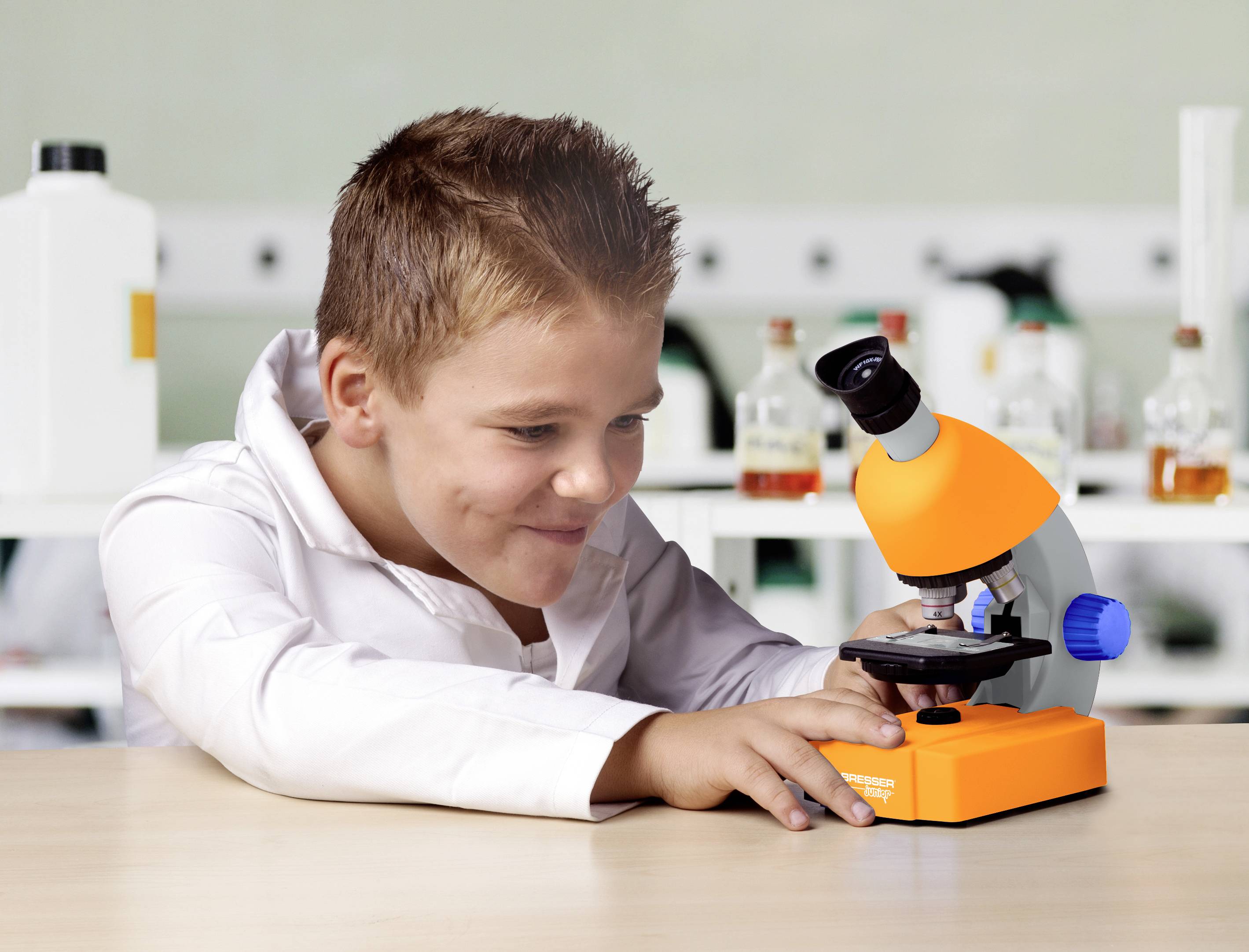 A child in a white shirt sits at a table and looks through a microscope. Laboratory equipment is visible in the background.