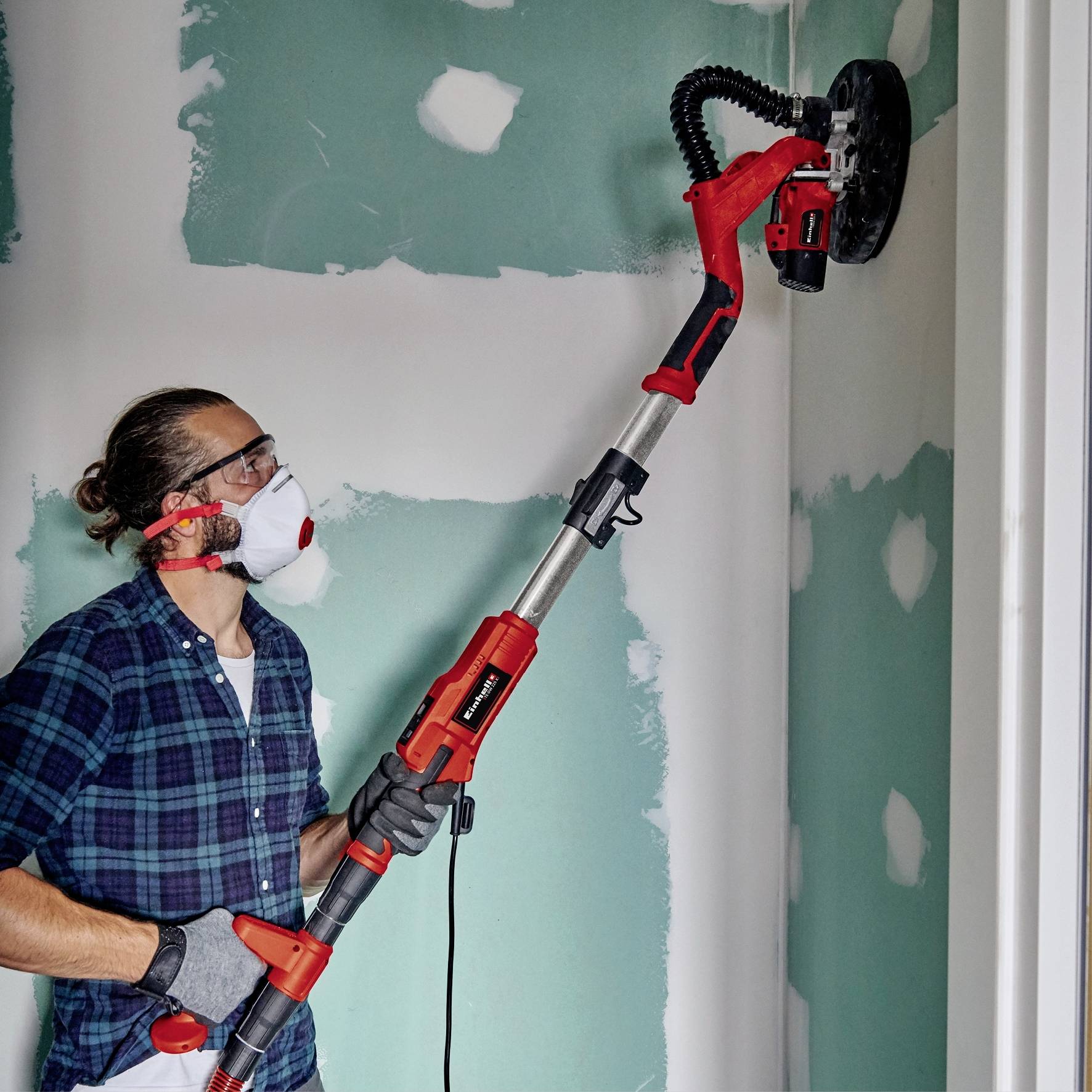 A person is sanding a wall with an electric sander. They are wearing safety glasses and a dust mask. Background: an unplastered wall.