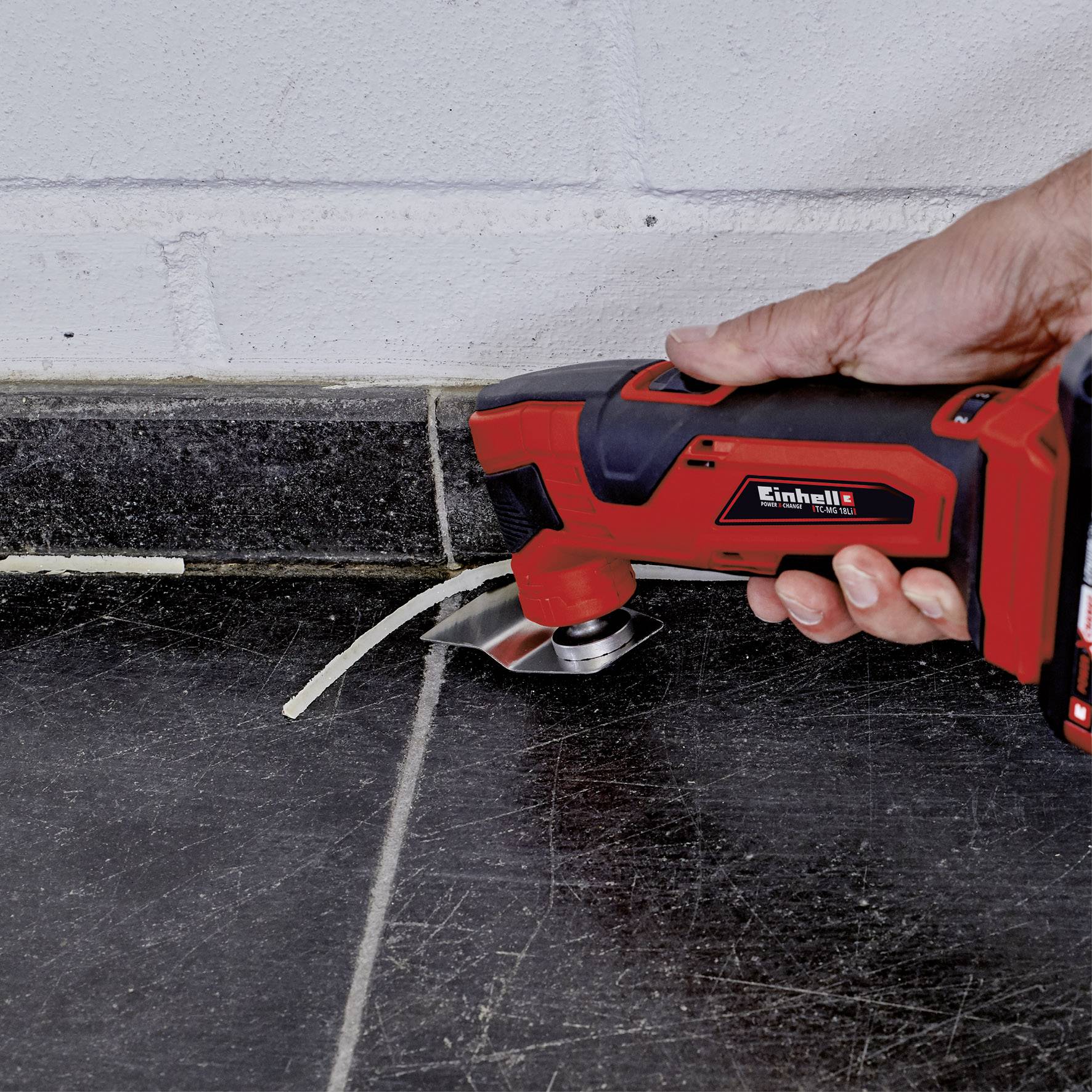 A hand holds a multi-tool cutting a fine line along a dark tiled floor next to a wall.