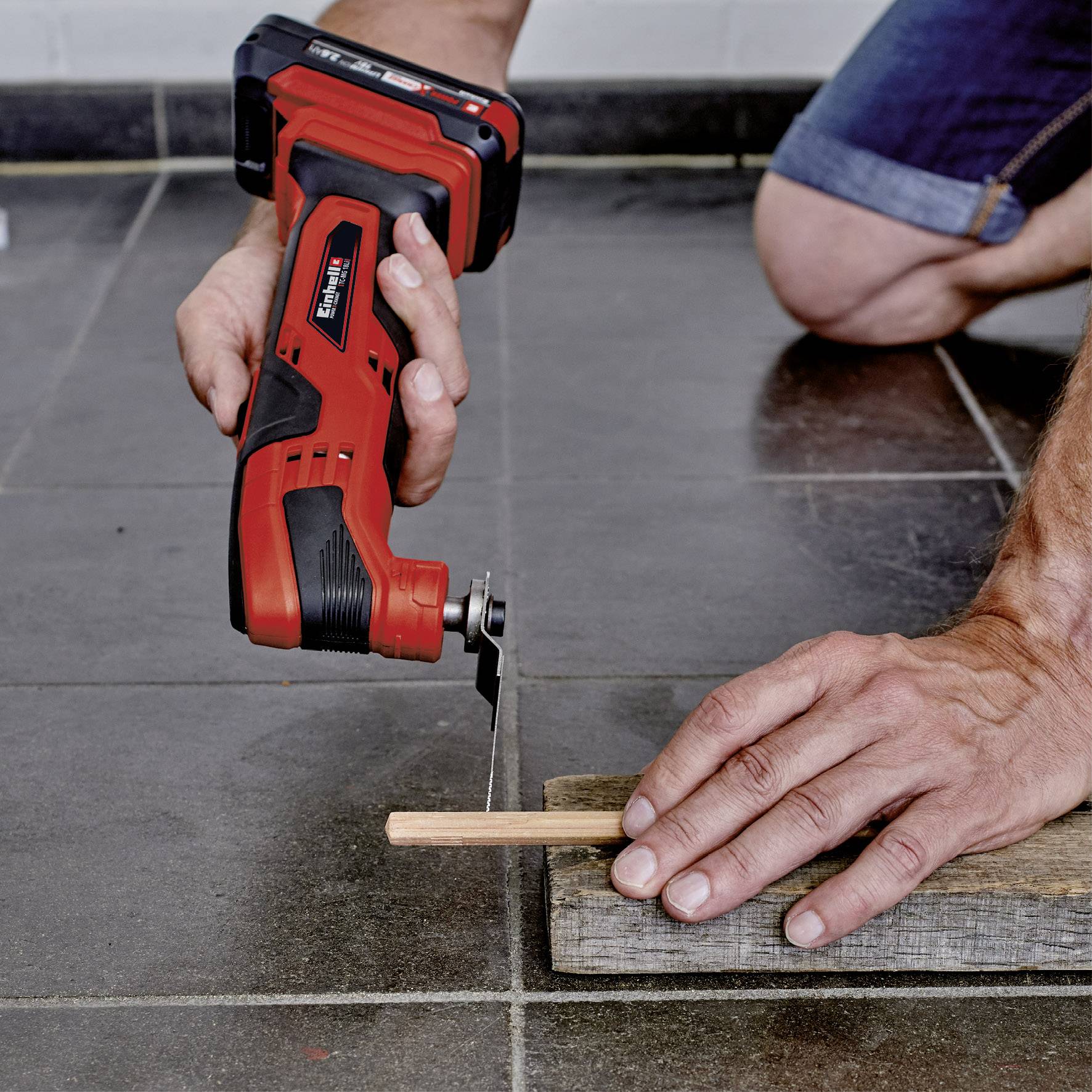 A person is using a red electric tool to cut a thin piece of wood. The wood is lying on a grey tiled floor.