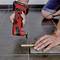 A person is using a red electric tool to cut a thin piece of wood. The wood is lying on a grey tiled floor.