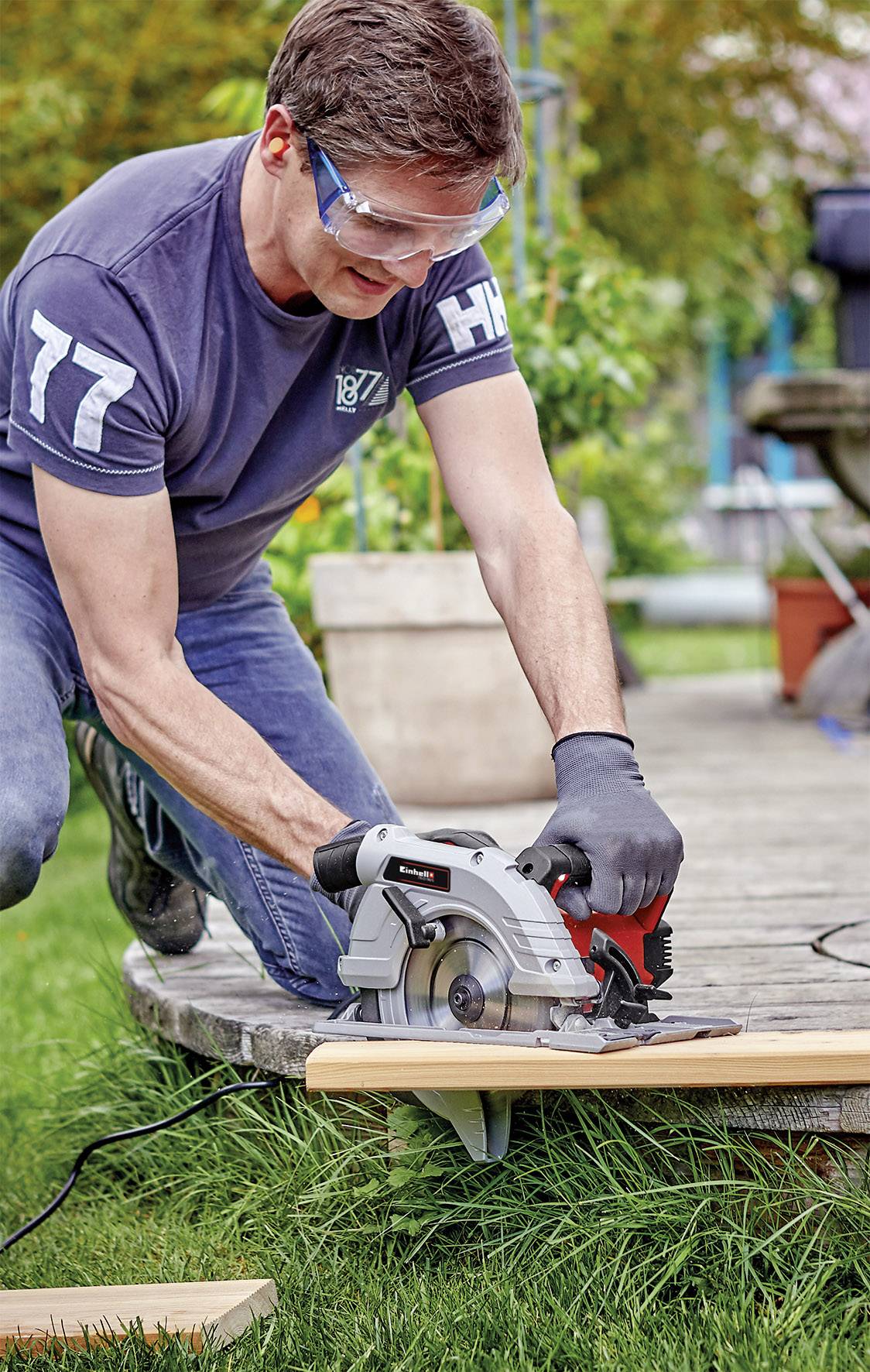 A man is cutting wood with a circular saw outdoors. He is wearing safety glasses and gloves. Plants and a terrace are visible in the background.
