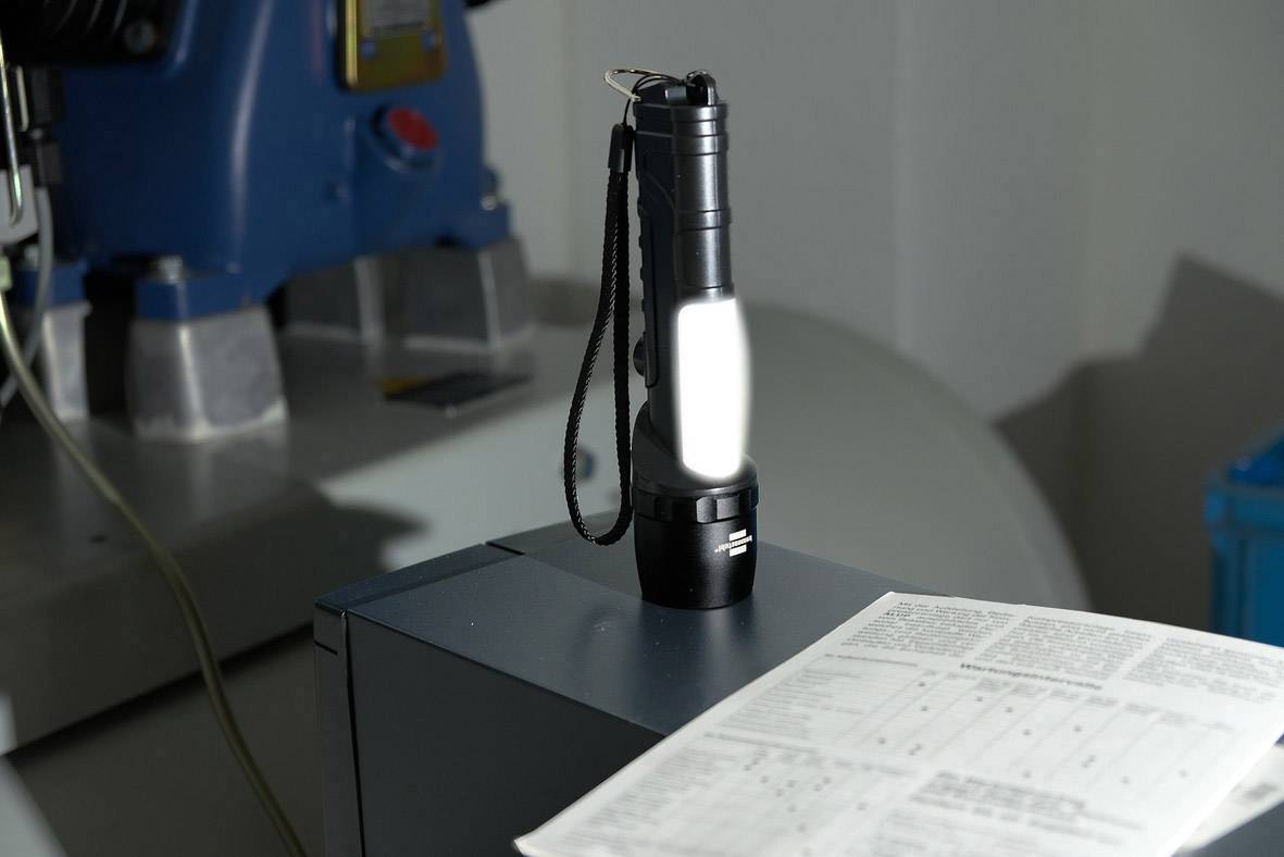 A black torch sits on a cupboard next to an open manual. Blurred machinery is visible in the background.