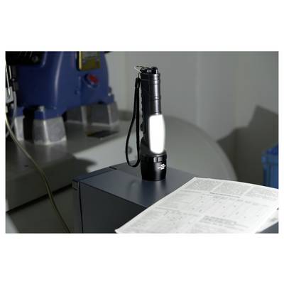 Black handheld inspection flashlight standing on a dark machine beside a printed instruction sheet in an industrial setting.
