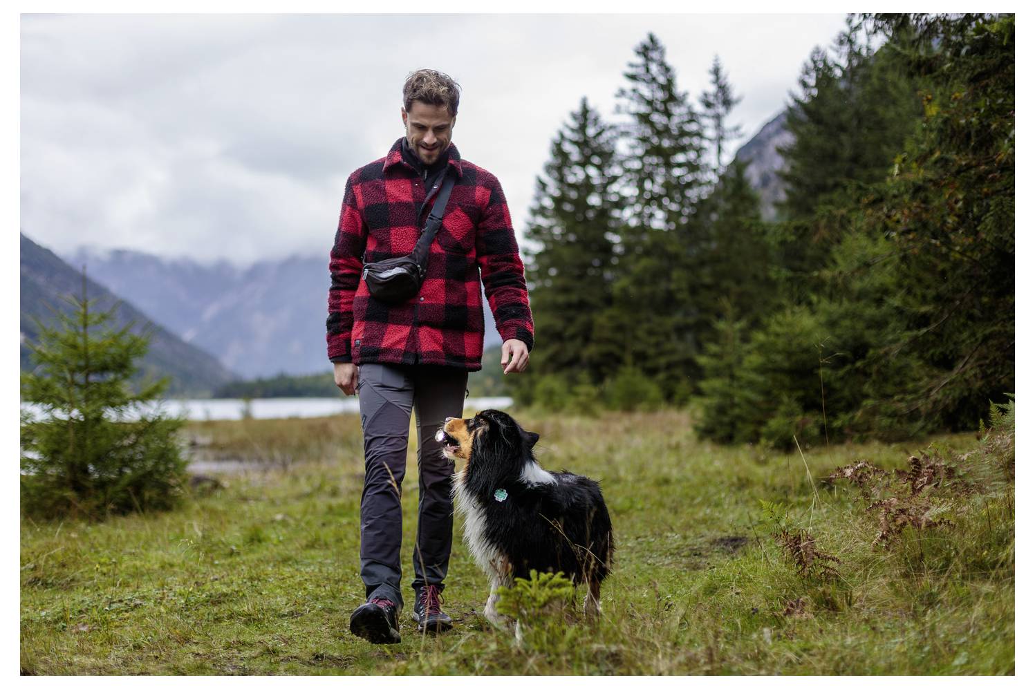 Man walks with a dog on a grassy trail near a lake and mountains, surrounded by evergreen trees on an overcast day.