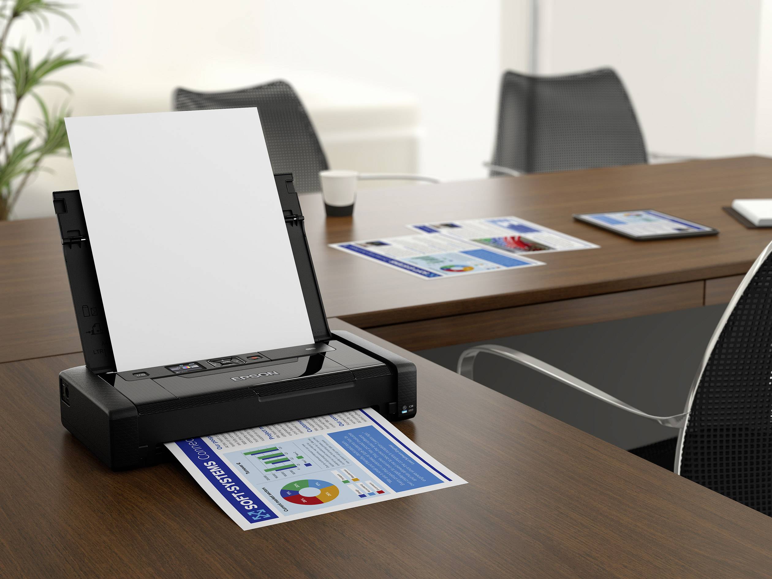 A printer sits on a desk in an office, printing a diagram. Blurred office chairs and other printouts are visible in the background.