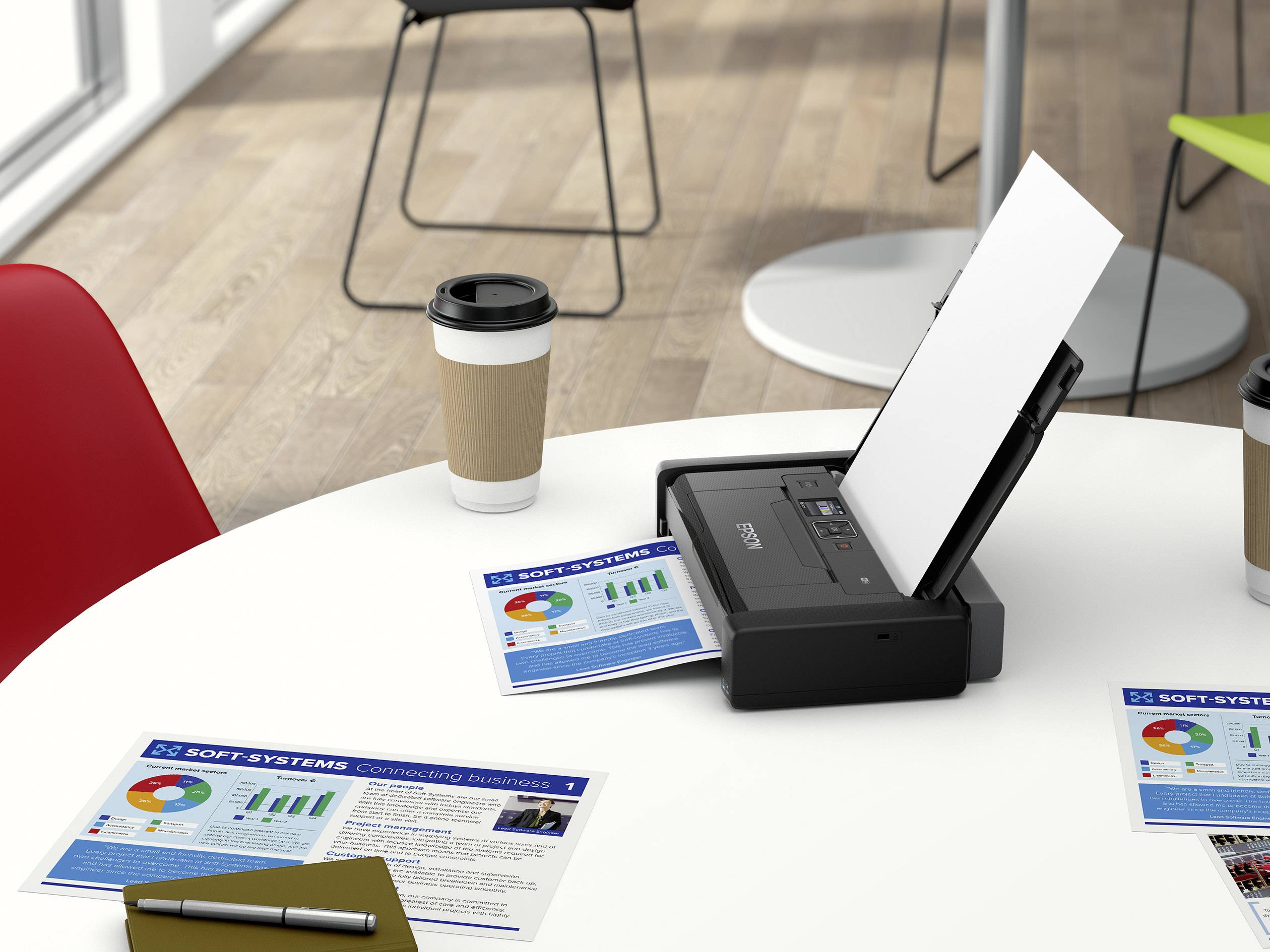 A scanner is lying on a table next to a coffee mug and two flyers with diagrams. Chairs are visible in the background.