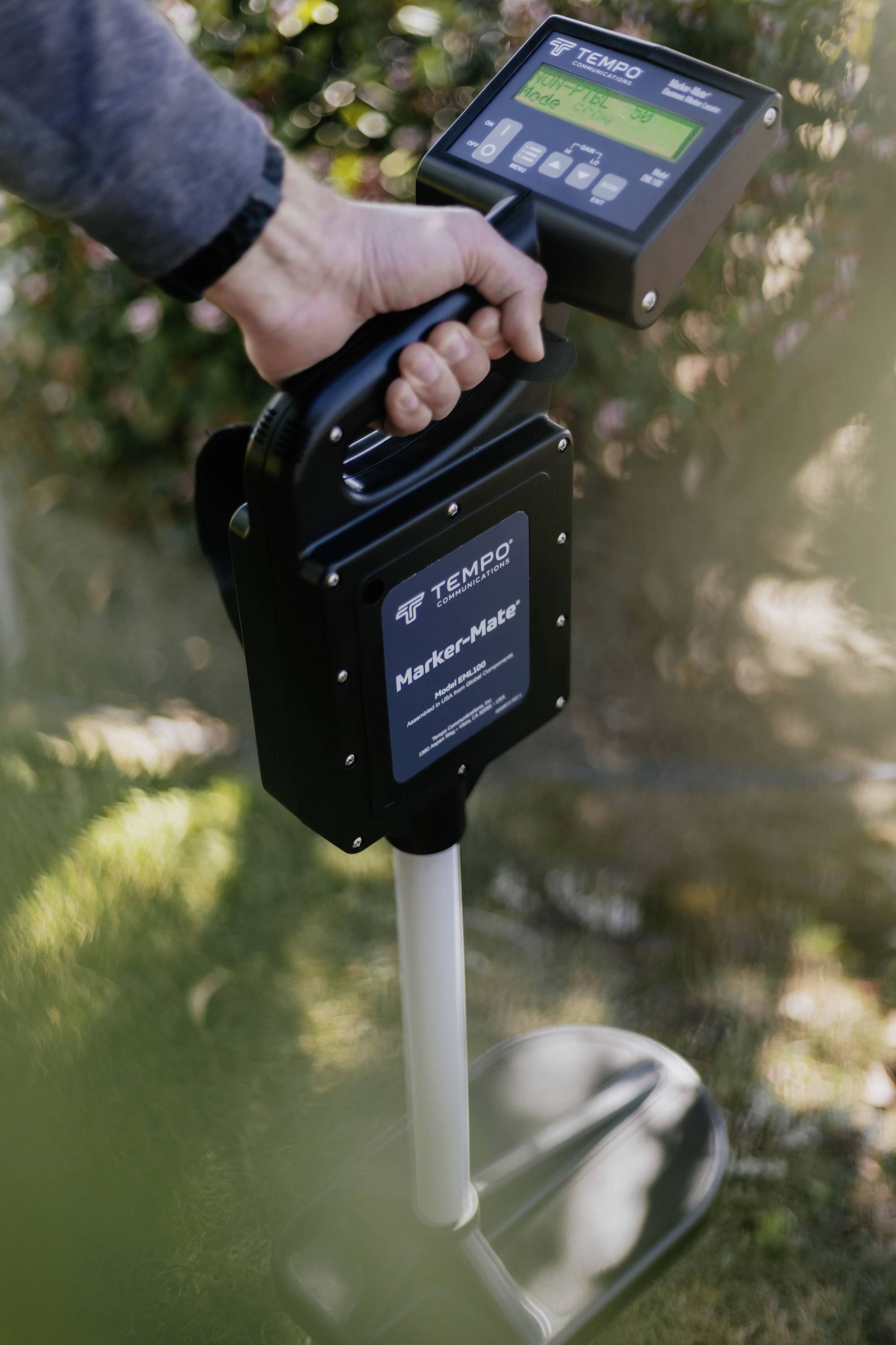 A person is holding a handheld device for detecting underground utilities, surrounded by grass and bushes.