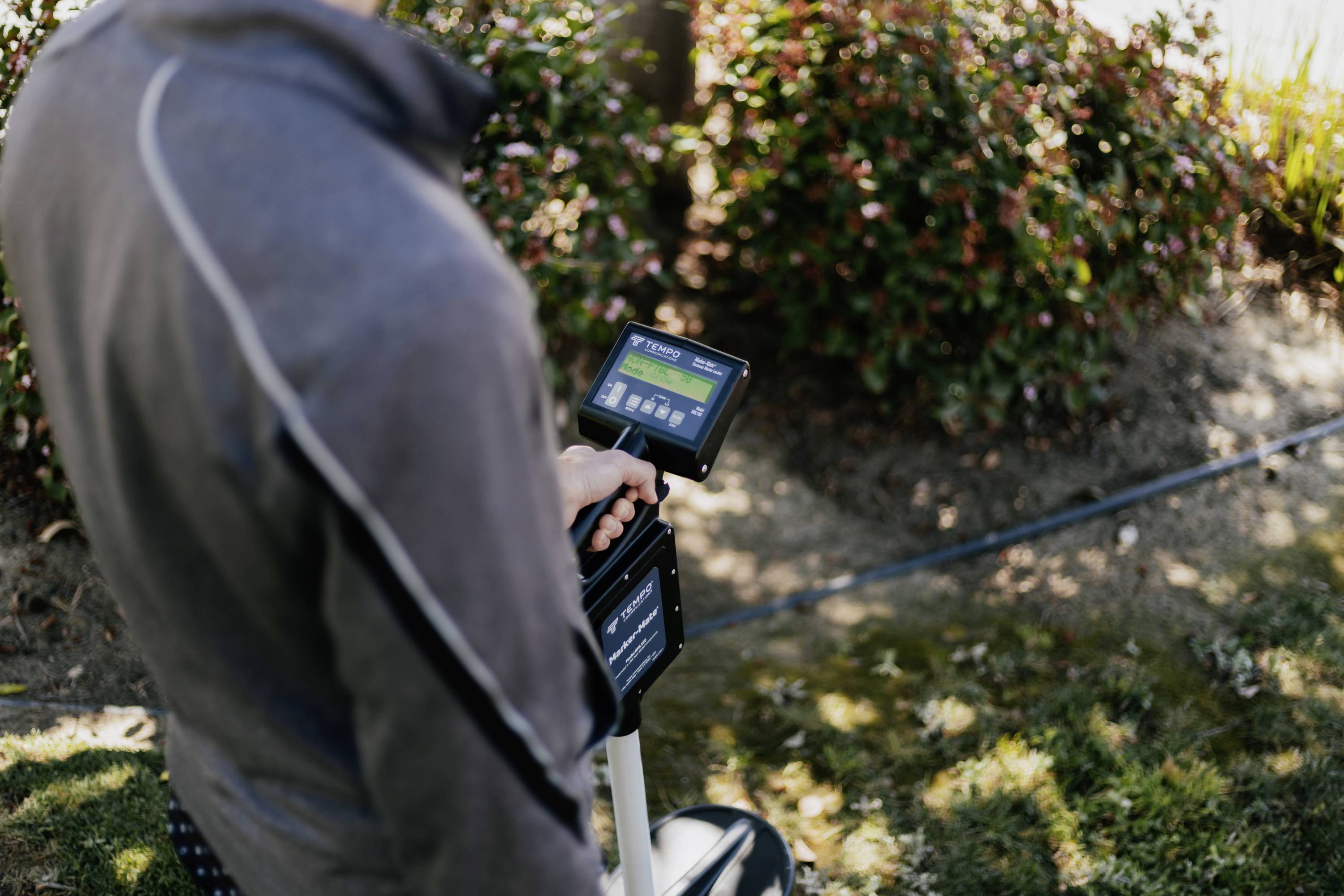 A person is using a portable device for outdoor measurements in a garden. The background shows bushes and plants.
