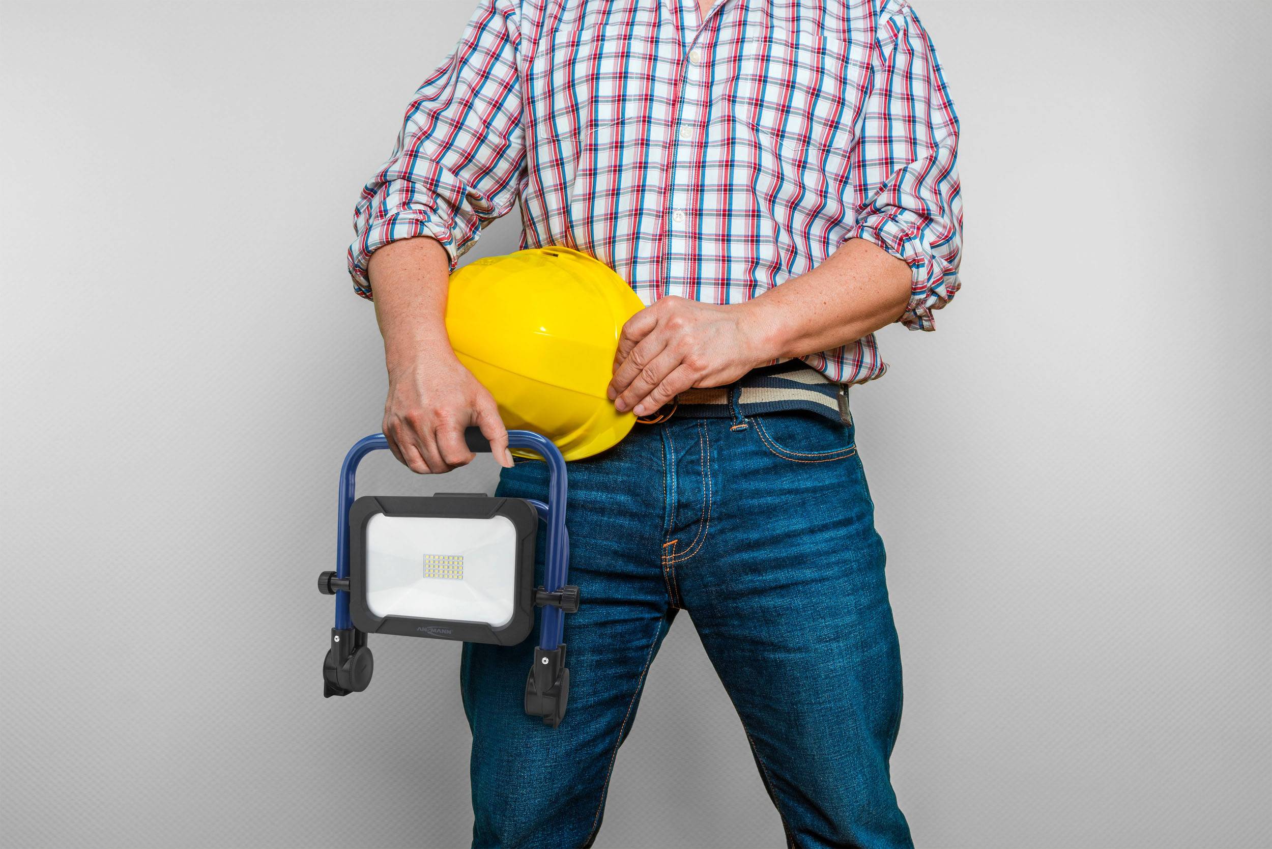 A person in a checked shirt is holding a yellow hard hat and a portable work light, standing against a grey background.