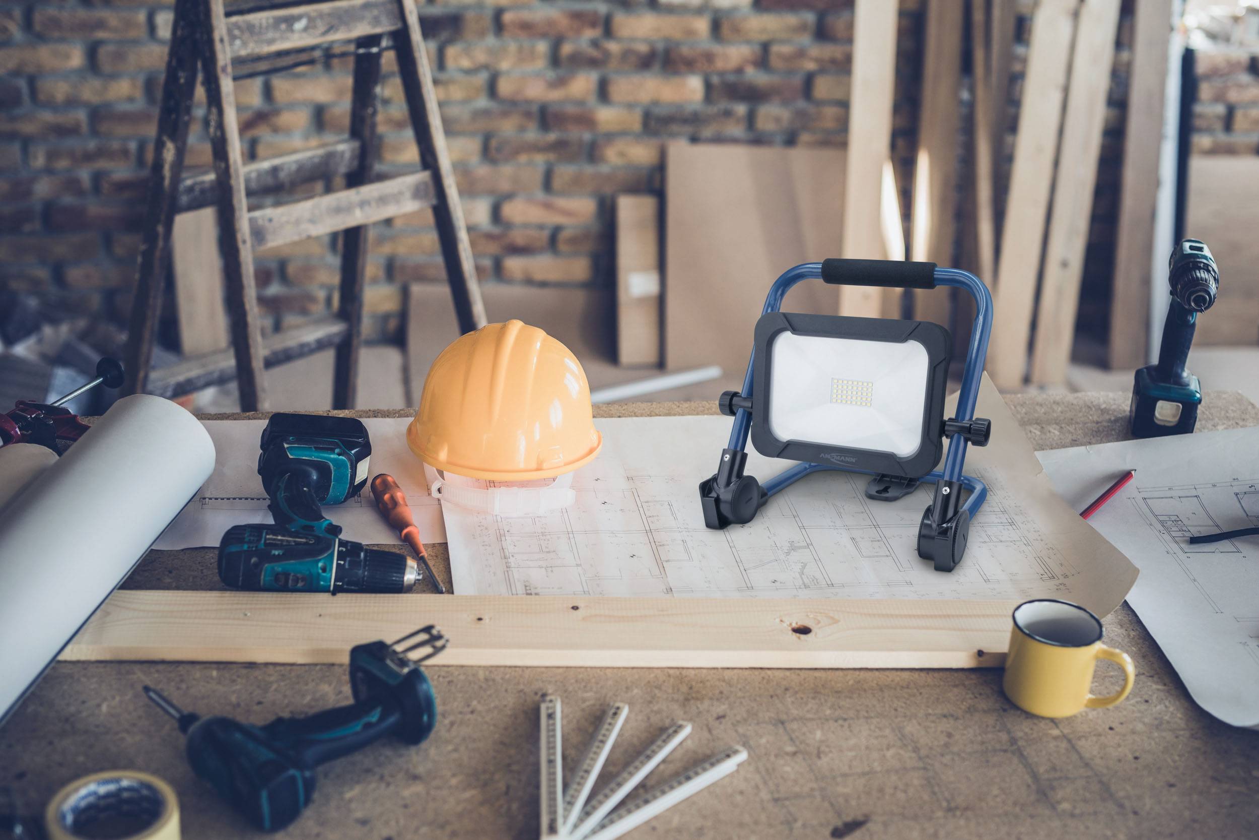 Workspace in a workshop with sketches, a hard hat, tools, a small lamp, and a coffee mug on a wooden table.