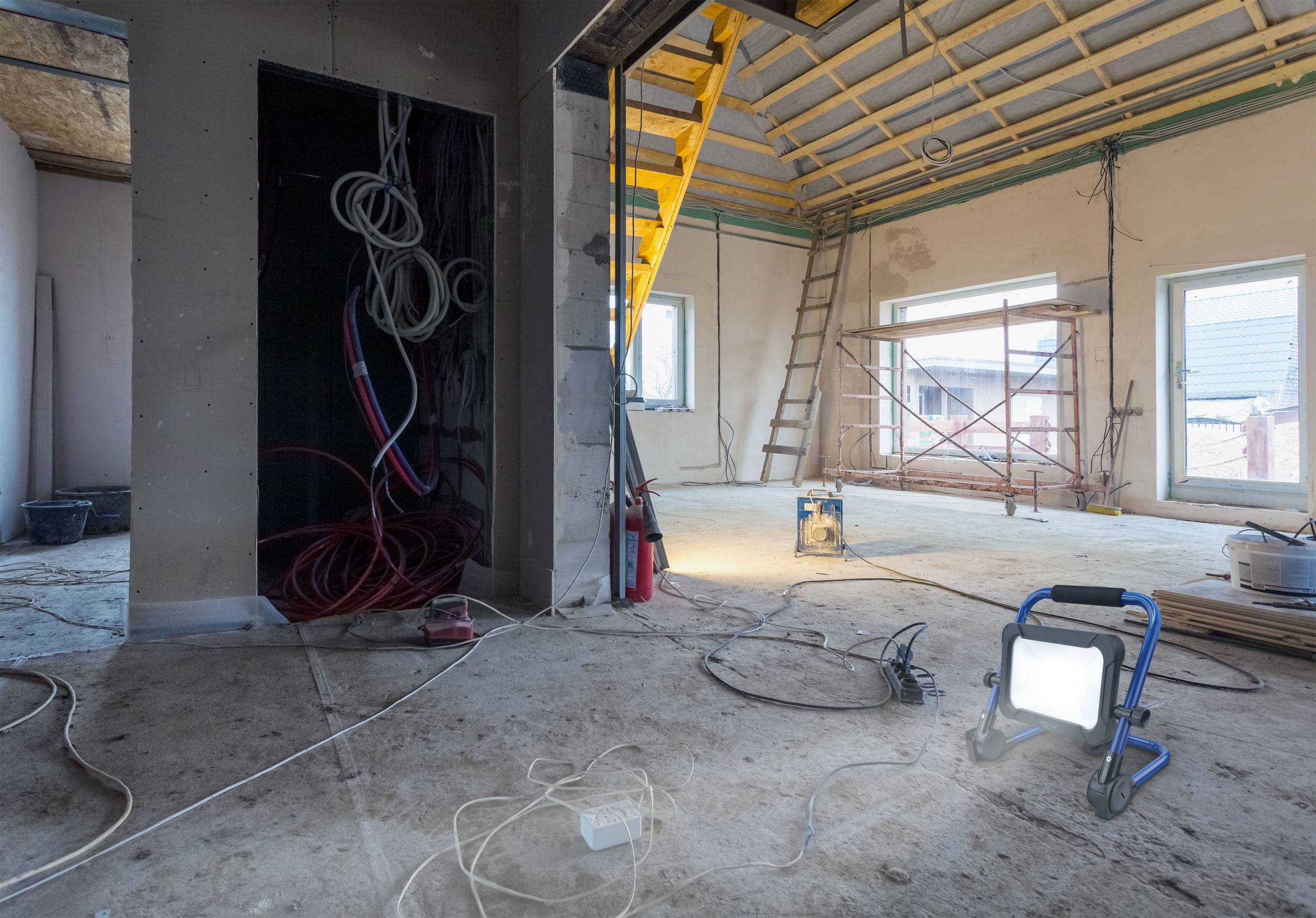 Construction site in a room with exposed roof trusses, cables, a ladder, and scaffolding. Daylight floods in through large windows.