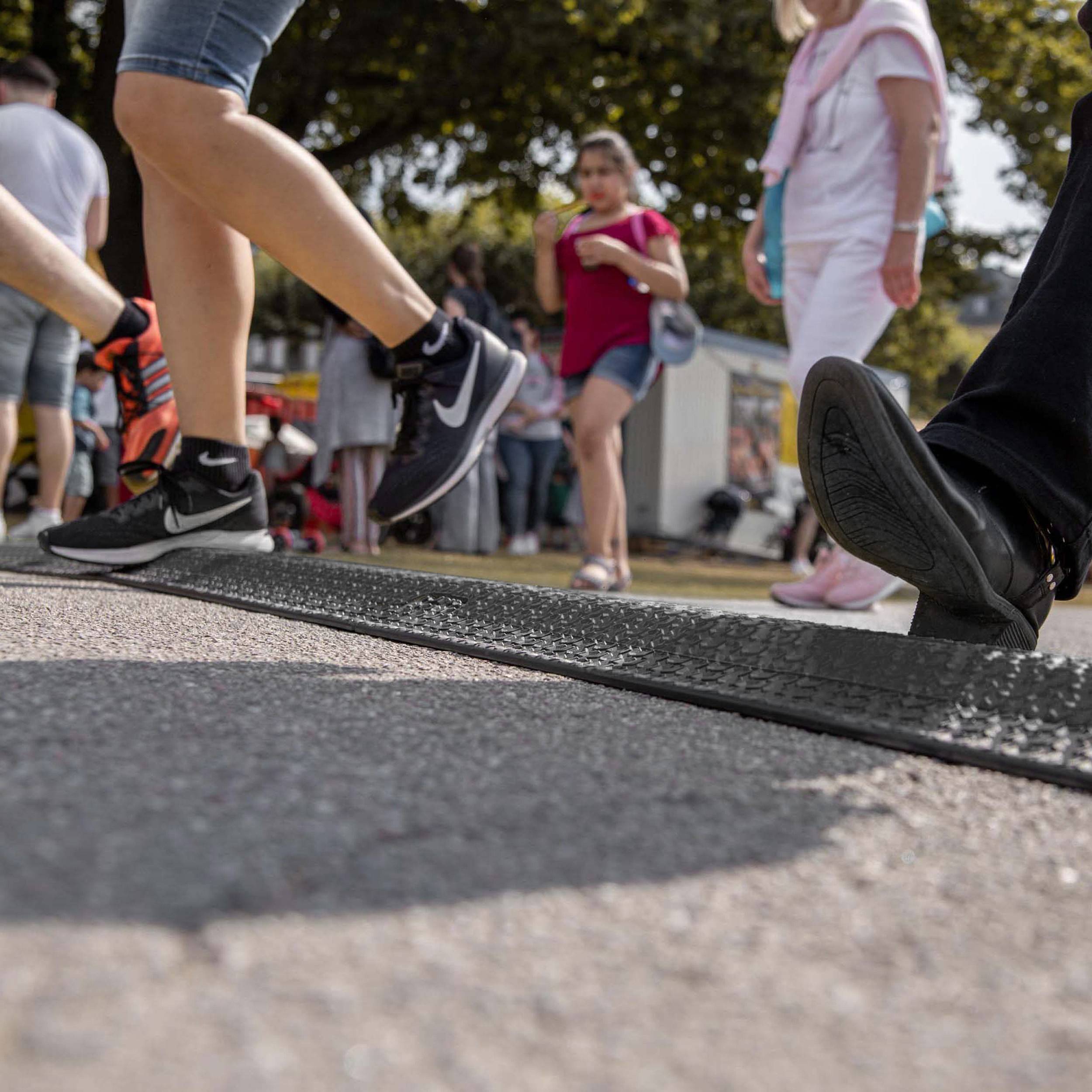 People are walking on a pavement. In the background, people are standing in front of a stall, some holding drinks in their hands.