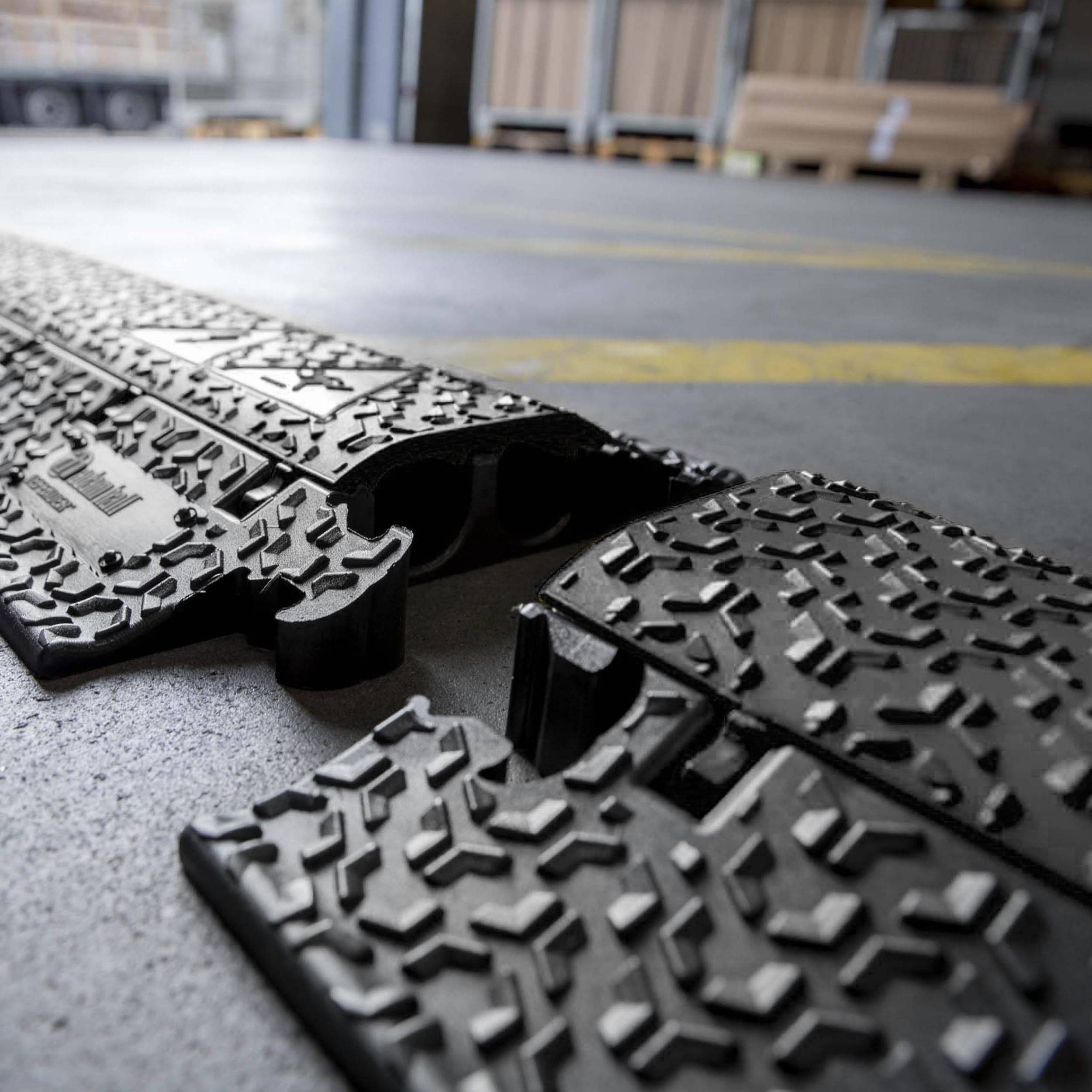 A cable trunking with a robust, non-slip design lies on a concrete floor in a warehouse environment. Cardboard boxes are blurrily visible in the background.