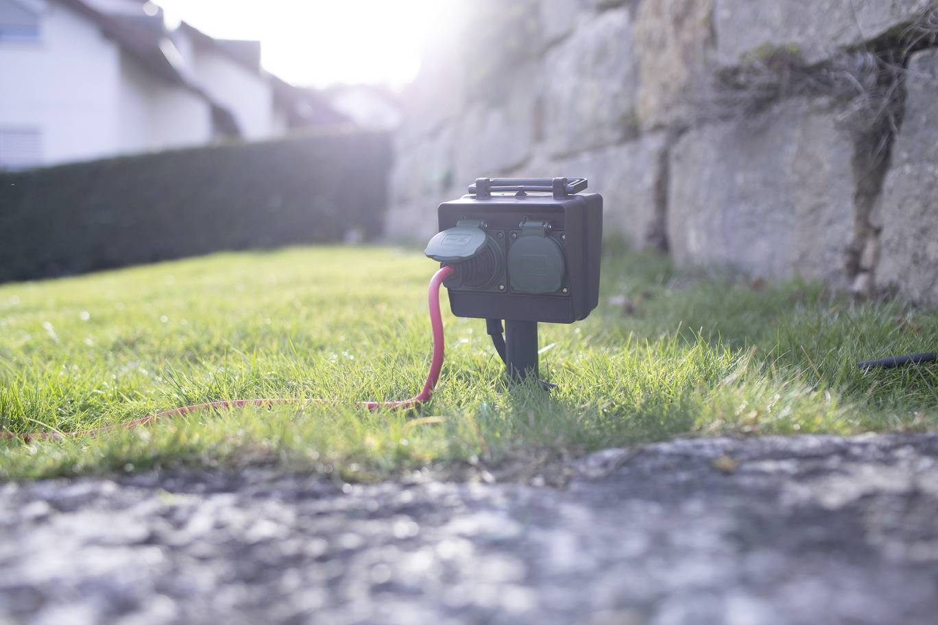 An outdoor socket with two connections in the garden, partially concealed by grass. A red cable is connected. Light is shining from behind.