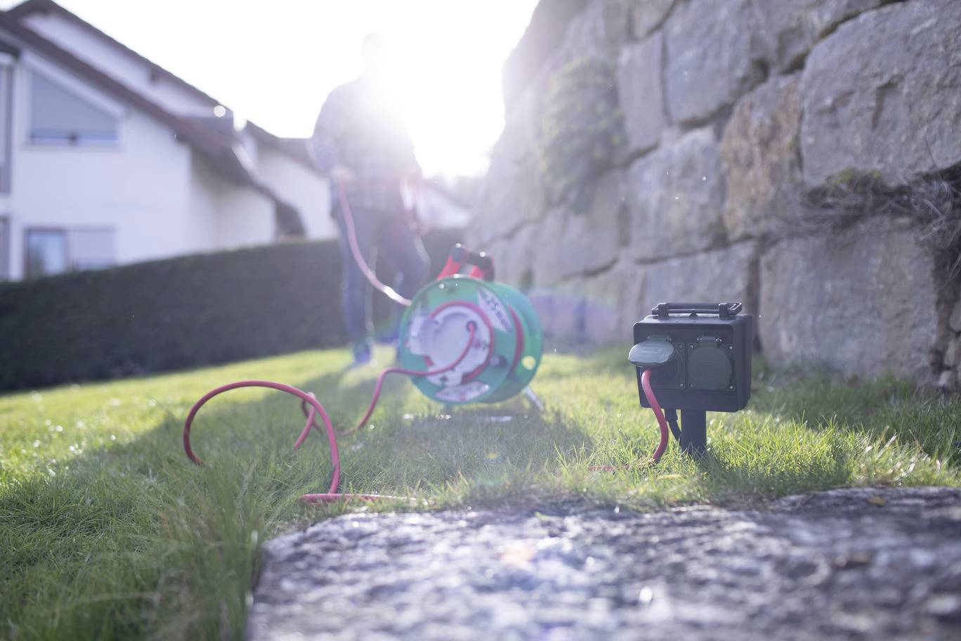 A man is pulling an extension lead across a lawn, near a house and a stone wall, on a sunny day.