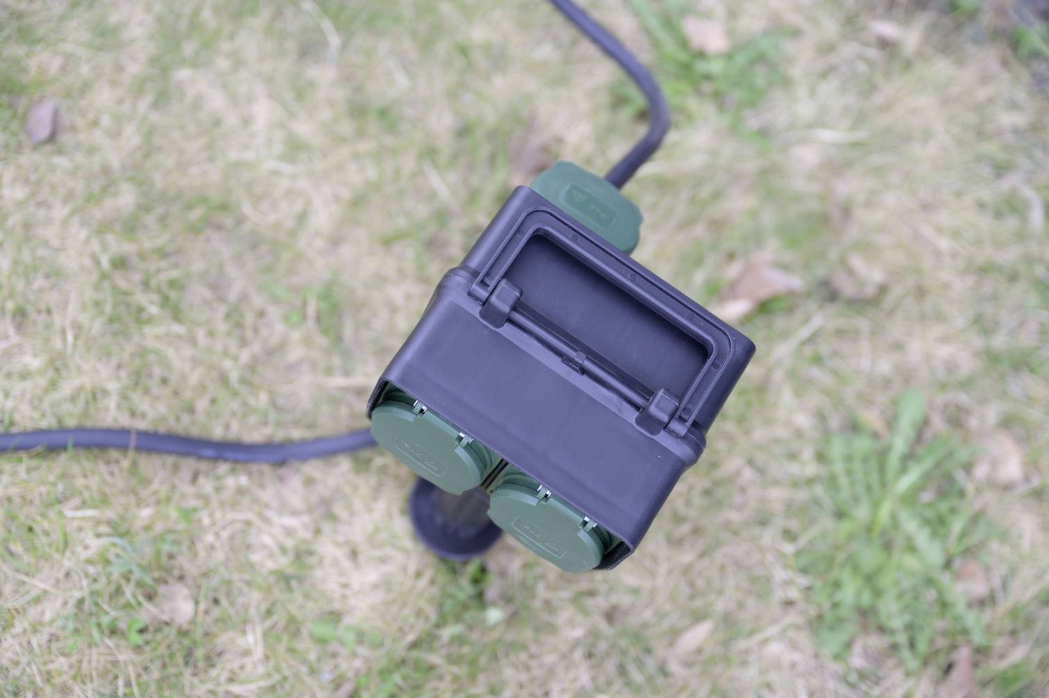 An outdoor socket with two green sockets and a black cable lies on a lawn.