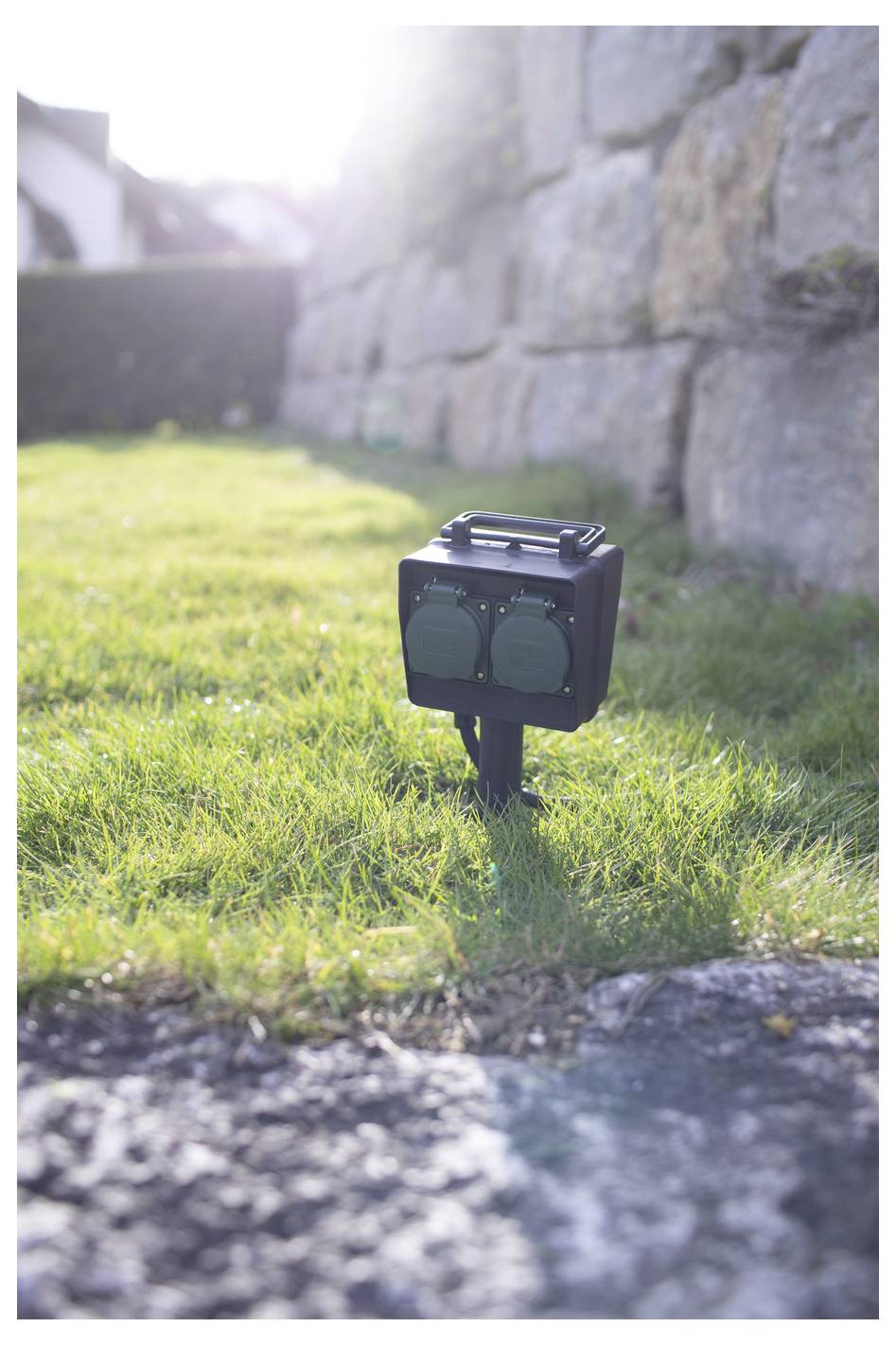 A small black outdoor electrical box on a stake sits in grass beside a stone wall, lit by bright sunlight.