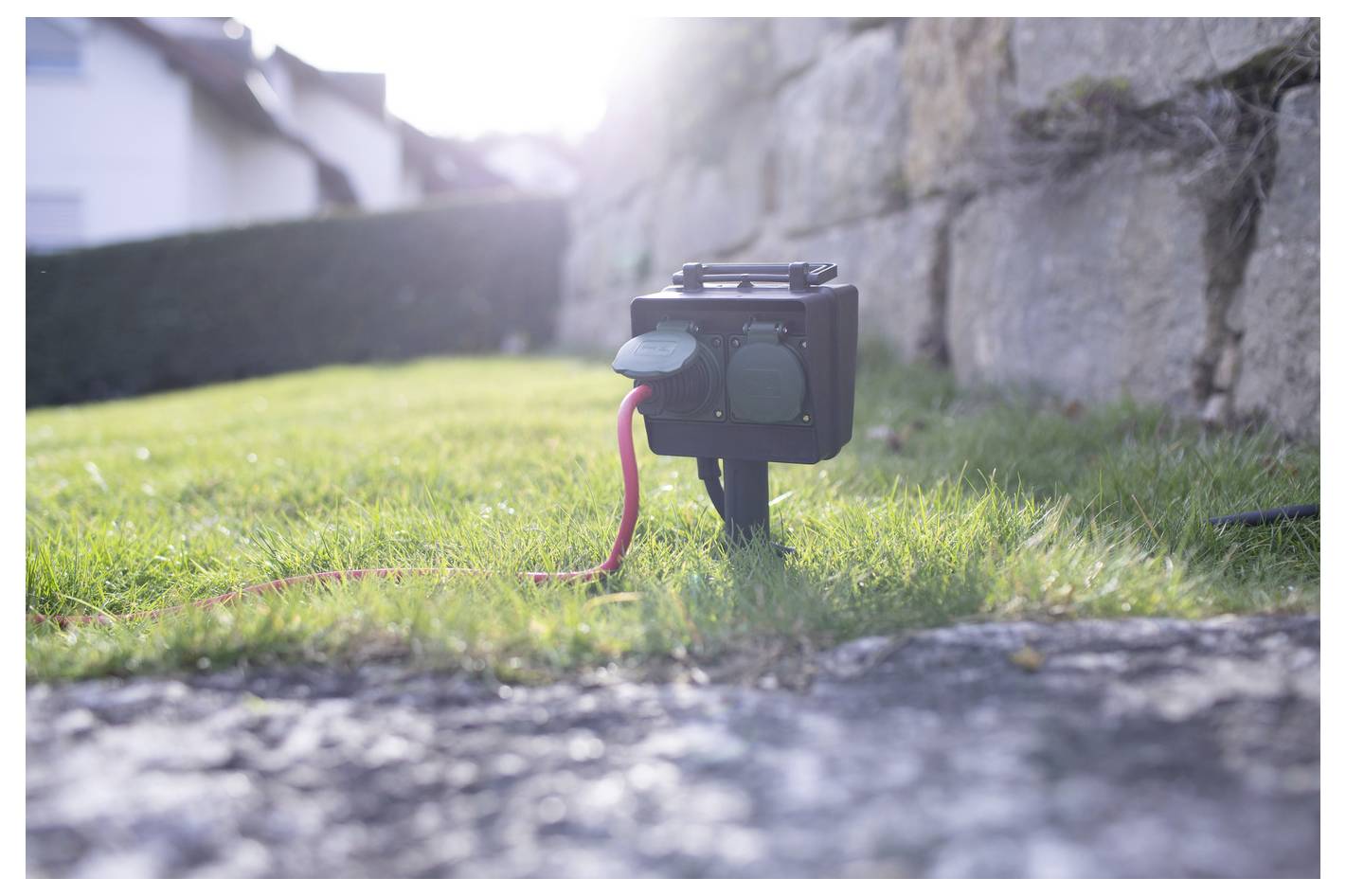 Small black action camera on a low tripod in sunlit grass, with a pink cable attached and a stone wall in the background.