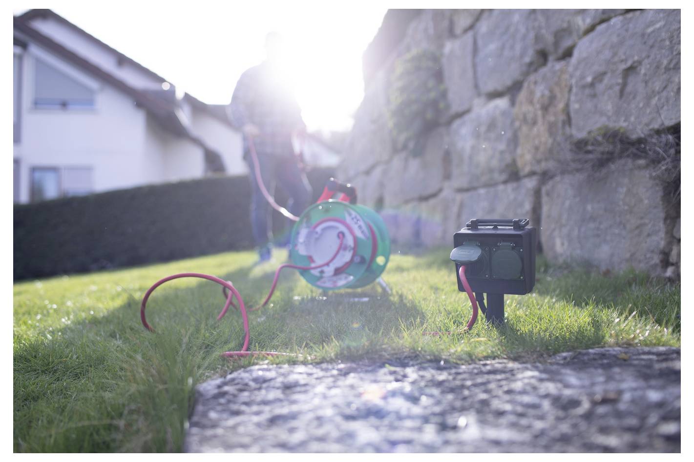 Person working outdoors with a green hose reel and a black pump or battery pack on the grass beside a stone wall in bright sunlight.