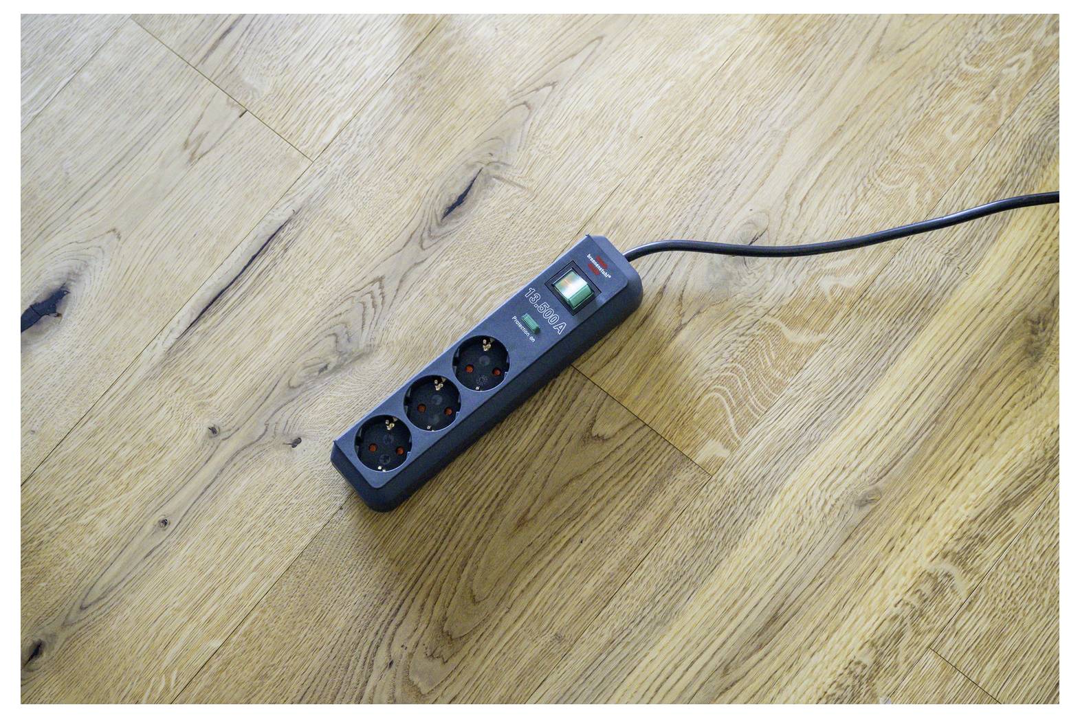 Black power strip with three outlets and a switch lying on a wooden floor, with its cord extending out of frame.