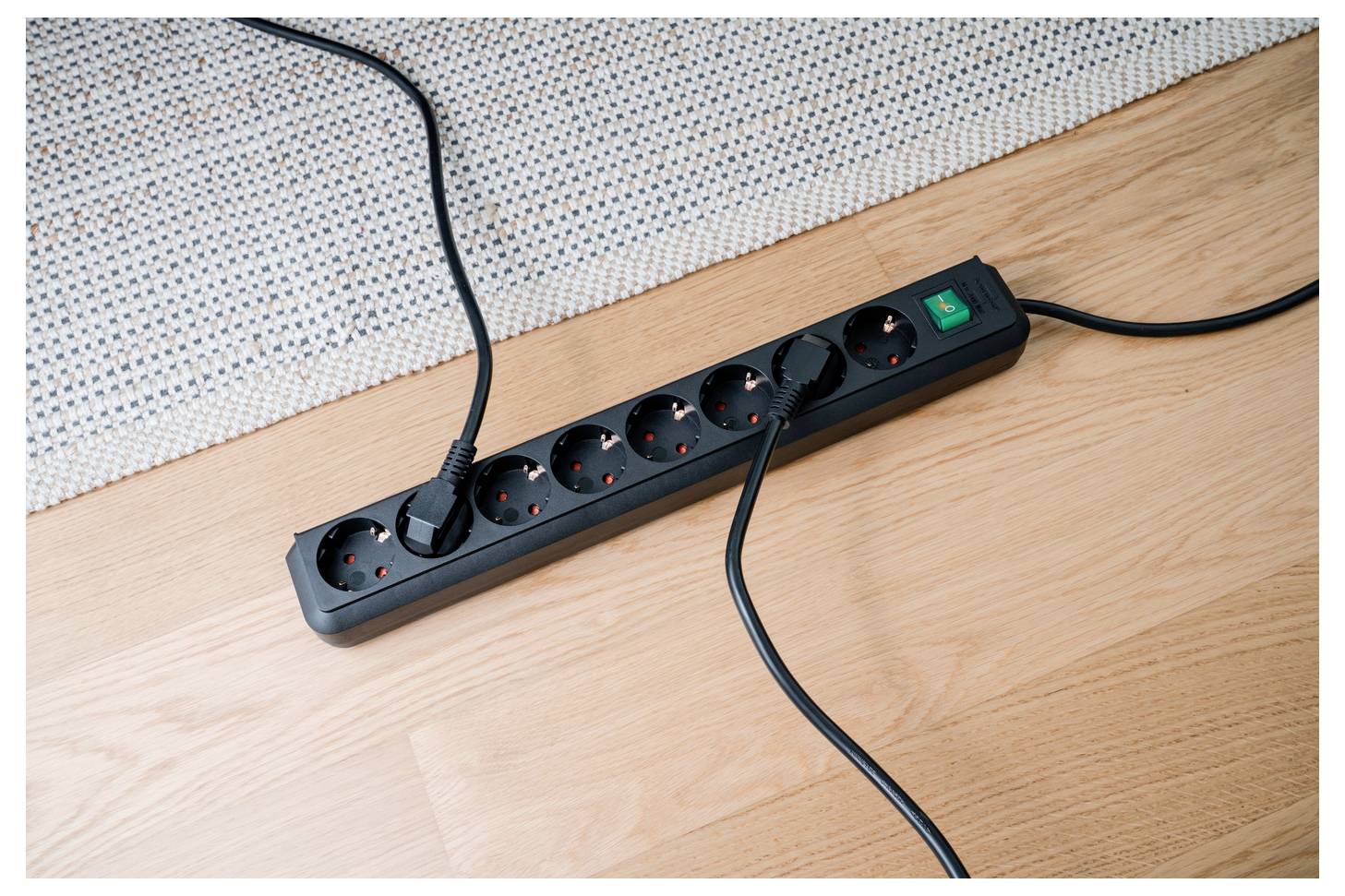 Black power strip with multiple plugged-in cords on a wooden floor, green on/off switch visible.<br>