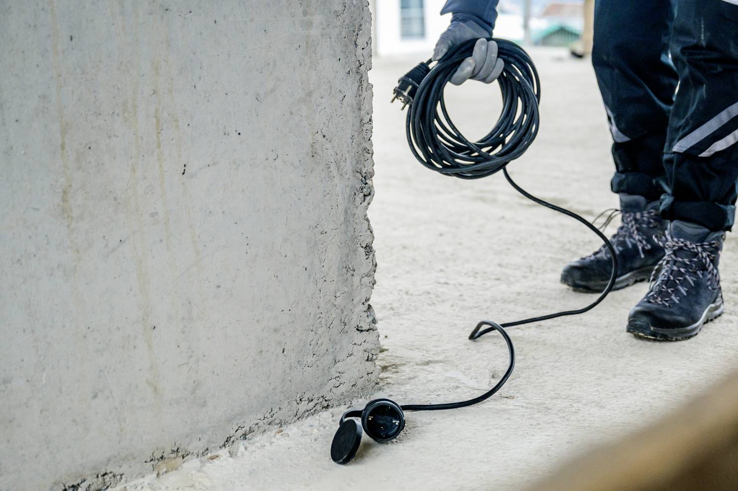 A person in protective clothing is holding a long, black extension lead near a concrete wall.