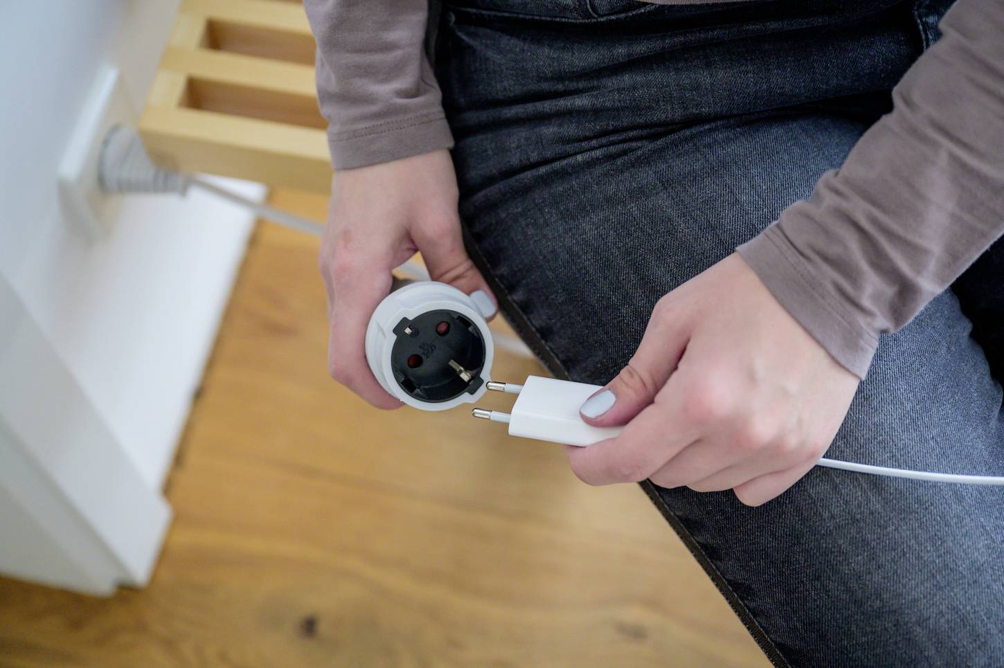 A person is plugging a white charging cable into a socket mounted on a wall. The background shows a wooden floor and the edge of a piece of furniture.