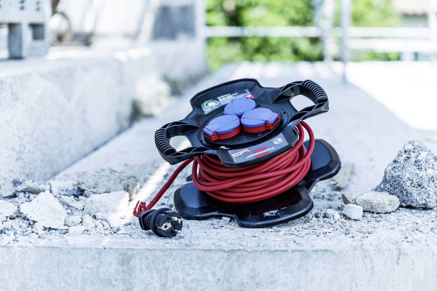 A black cable reel with a red cable and multiple sockets lies on a concrete floor, surrounded by rubble on a construction site.